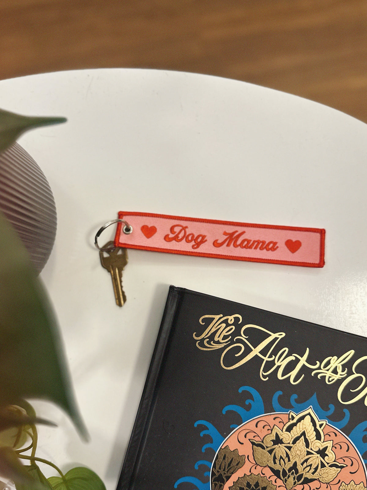 Pink and red keychain with 'Dog Mama' tag on a book titled 'The Art of...' on a white surface.