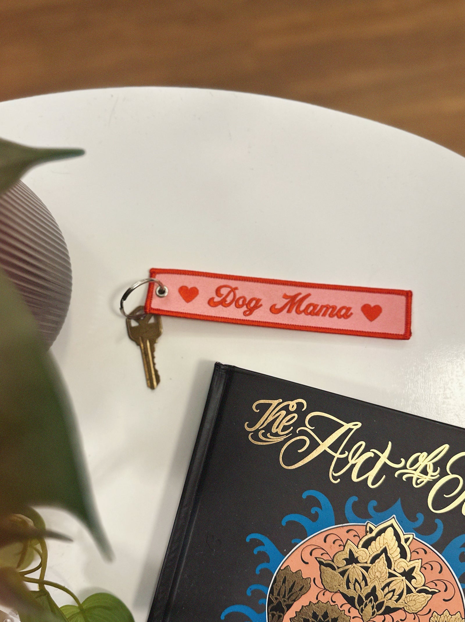 Pink and red keychain with 'Dog Mama' tag on a book titled 'The Art of...' on a white surface.