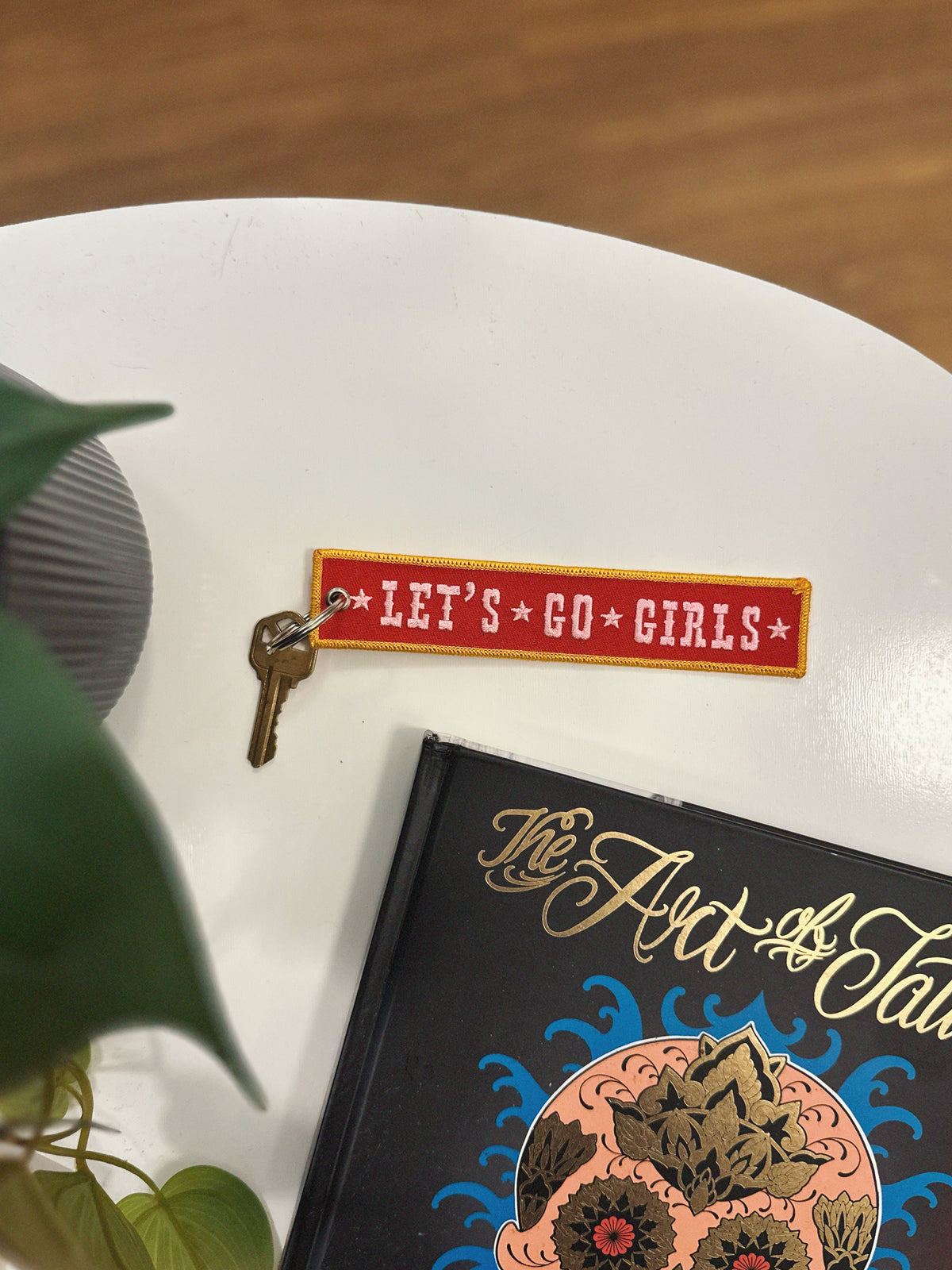 Embroidered red, pink, and yellow keychain with 'Let's Go Girls' on a white table with a plant and The Art of Tattoo in the background