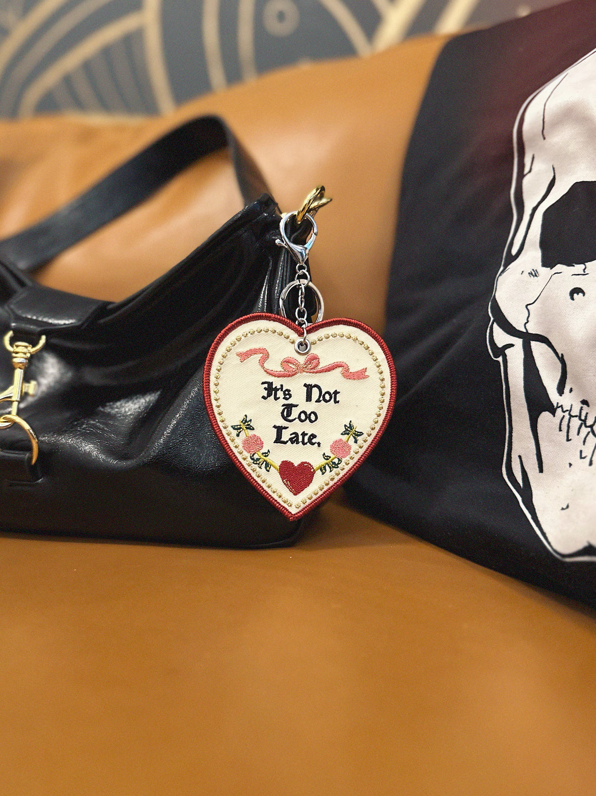 Heart-shaped keychain with 'It's Not Too Late' text attached to a black handbag.