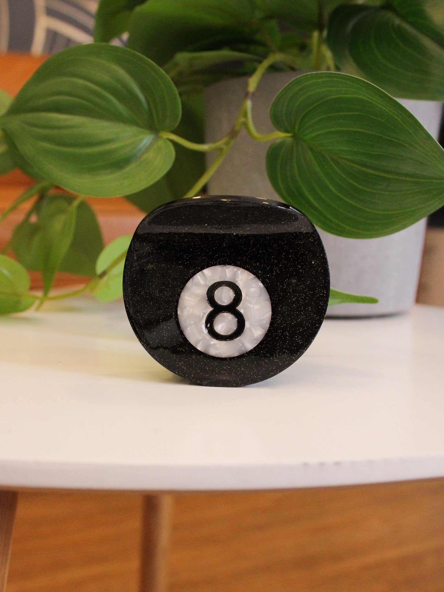 Black eight-sided die with number 8 on a white surface with green plants in the background