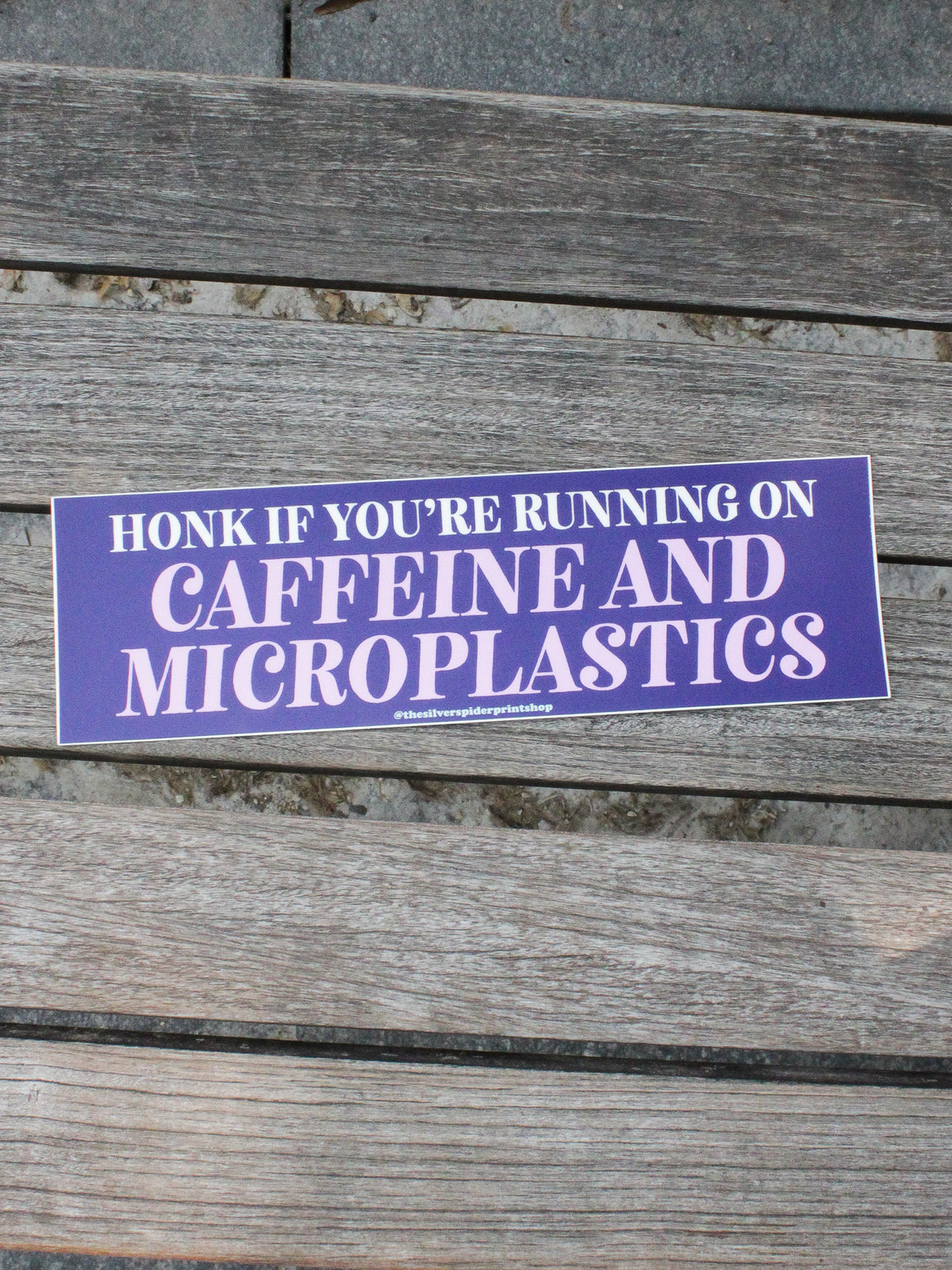 Purple sticker with text &#39;Honk if you&#39;re running on caffeine and microplastics&#39; on a wooden surface