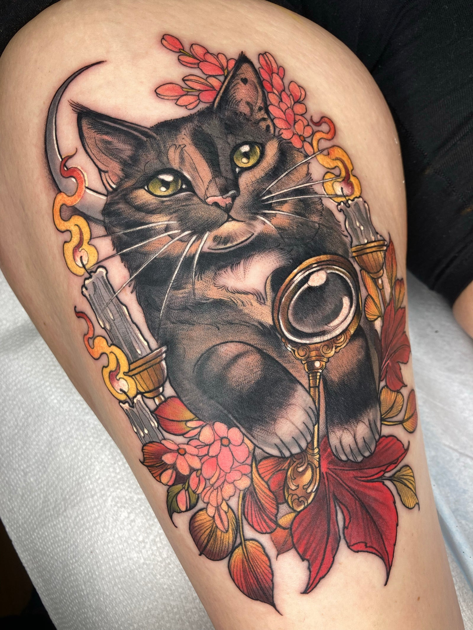 Neo-traditional cat portrait tattoo. This thigh tattoo in a warm color palette features red flowers, lit candles, and an ornate gold magnifying glass.