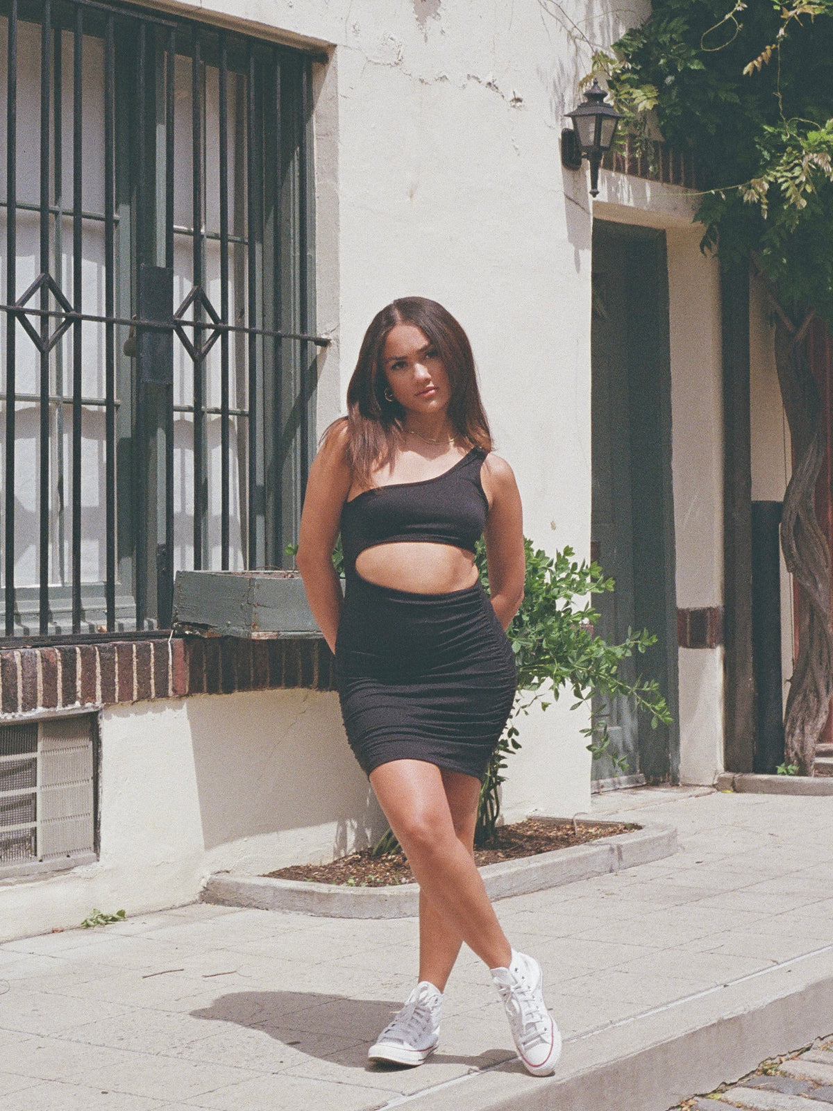 A woman wearing a one shoulder cut out mini dress with a ruched front in black color.