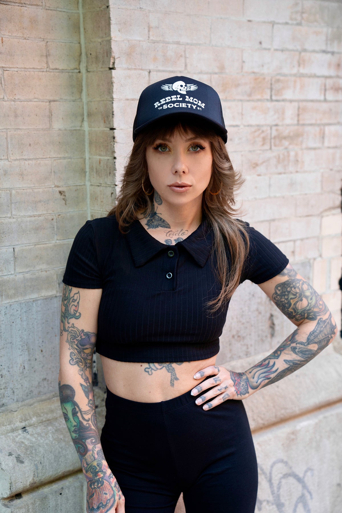 Megan Massacre wearing an alternative, rock n roll style black trucker hat which reads &quot;Rebel Mom Society&quot; in white with a skull logo. 