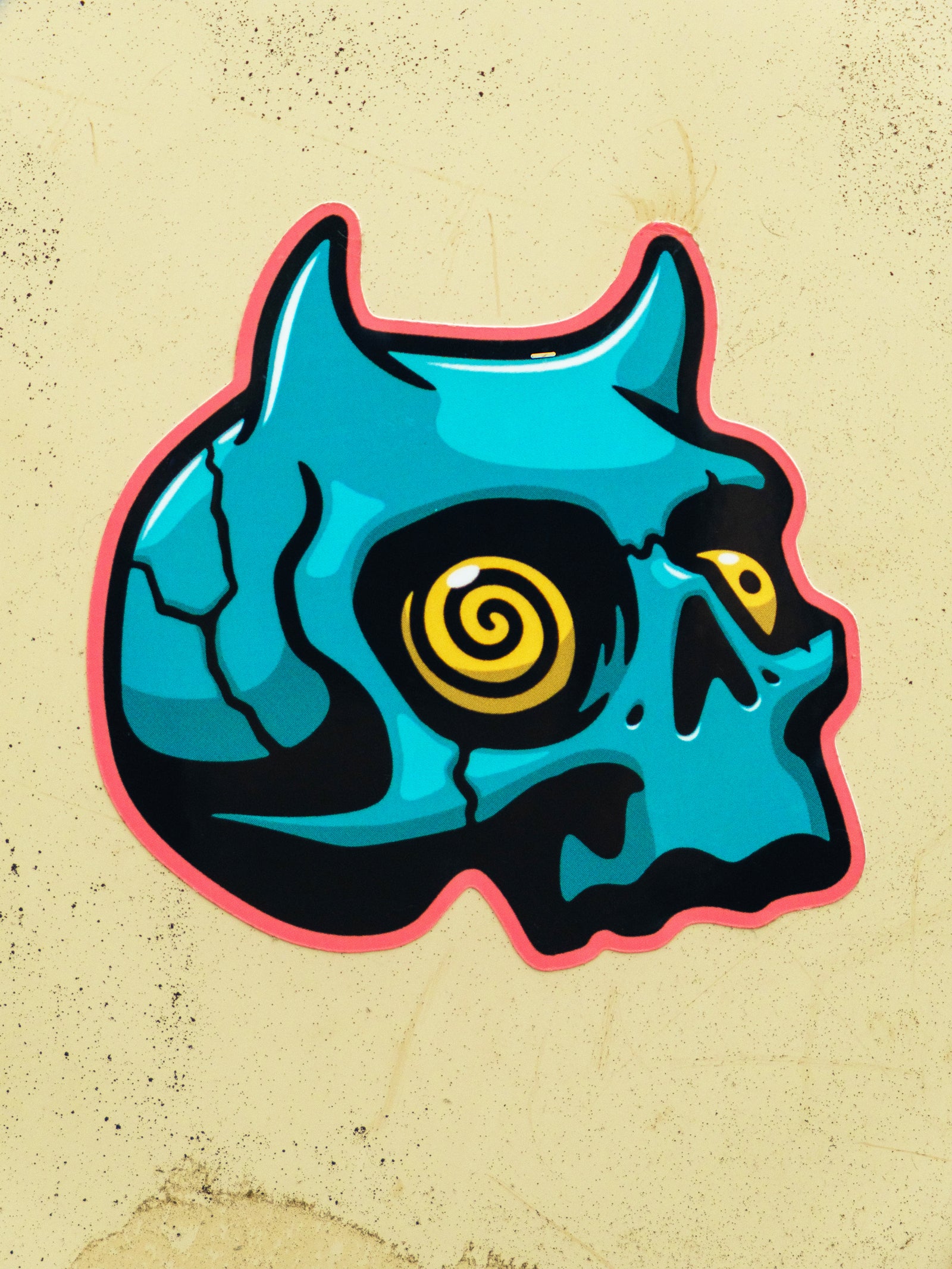 A die cut sticker of a teal-colored demon skull with a spiral yellow eye, designed by Cavan Infante.