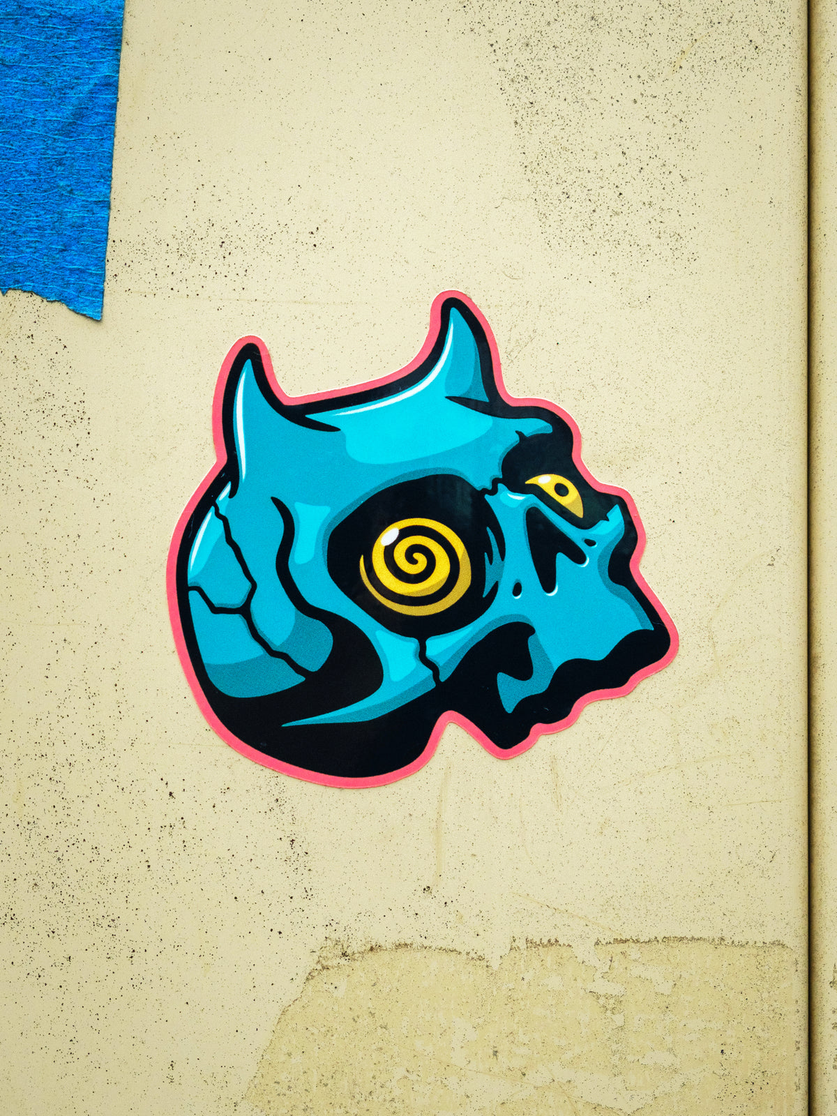 A die cut sticker of a teal-colored demon skull with a spiral yellow eye, designed by Cavan Infante.