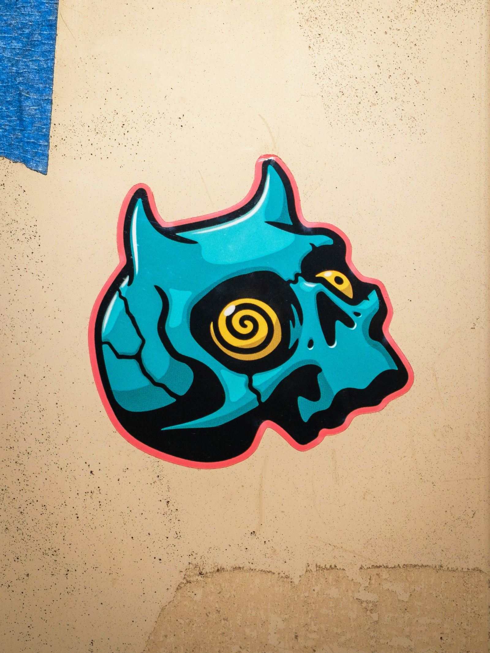 A die cut sticker of a teal-colored demon skull with a spiral yellow eye, designed by Cavan Infante.