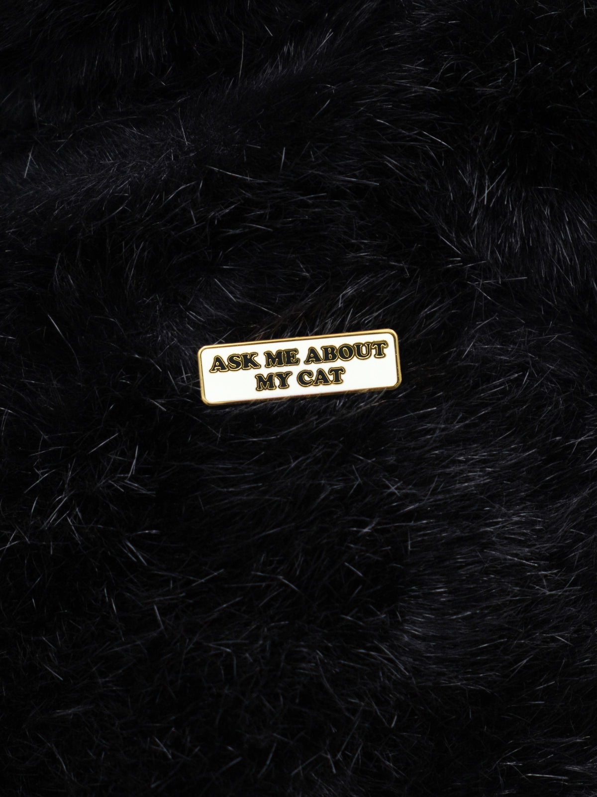 An enamel pin with the text 'ASK ME ABOUT MY CAT' attached to a black fuzzy background.
