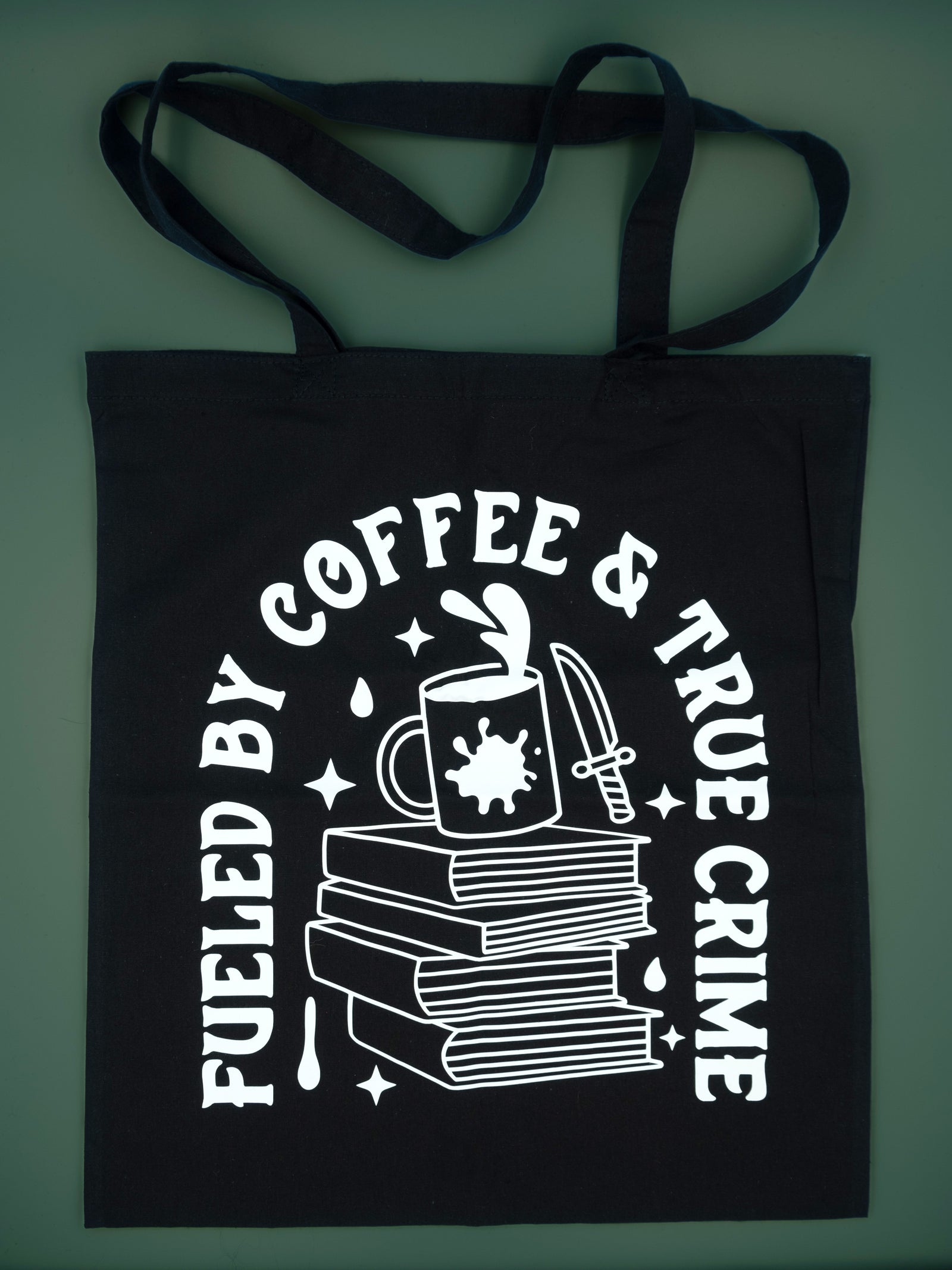 Black canvas totebag on a green background, tote bag reads Fueled By Coffee & True Crime