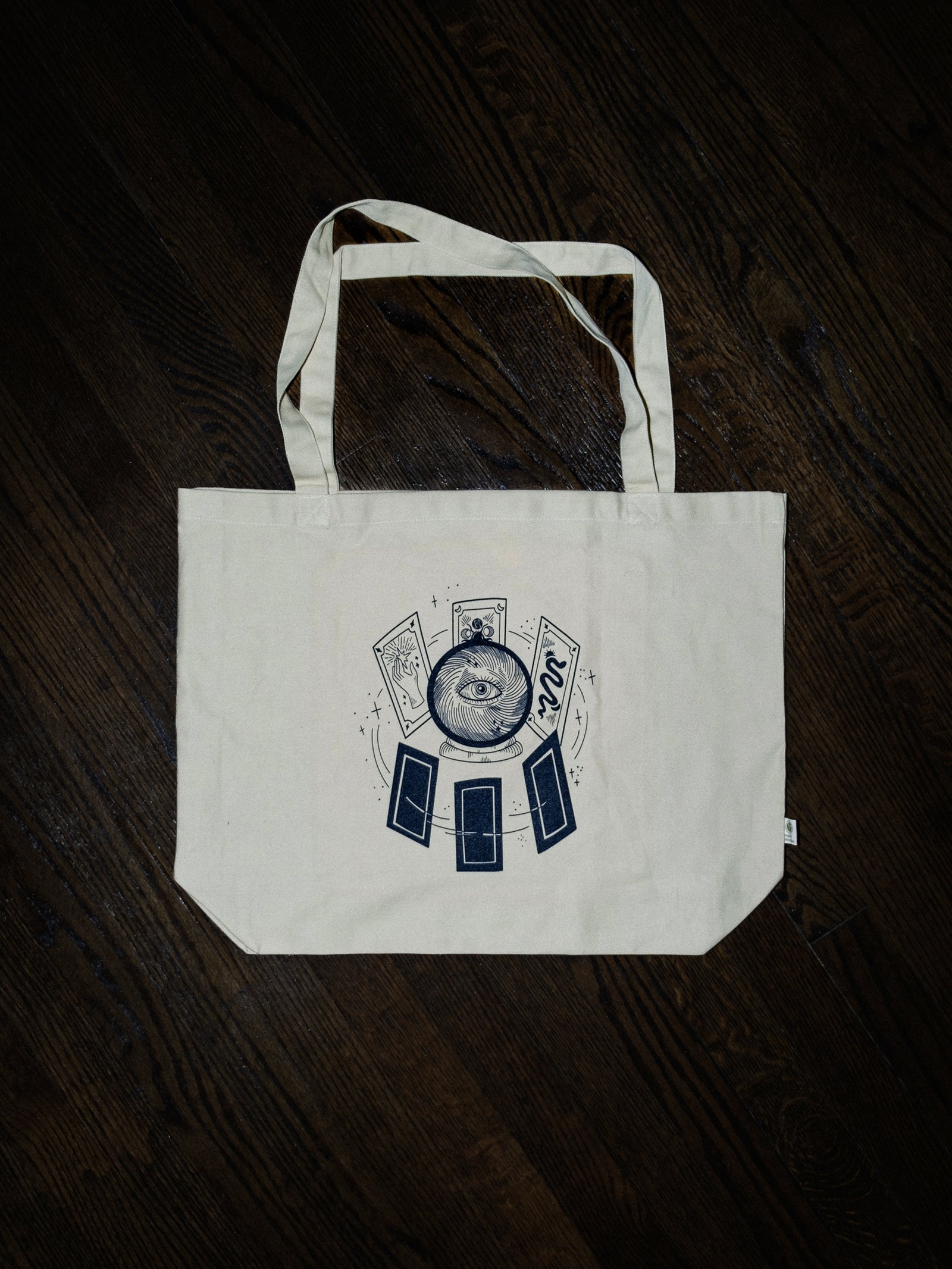 A white organic cotton tote bag with dual straps and a black graphic of a crystal ball and tarot cards on the front, placed on a dark surface.