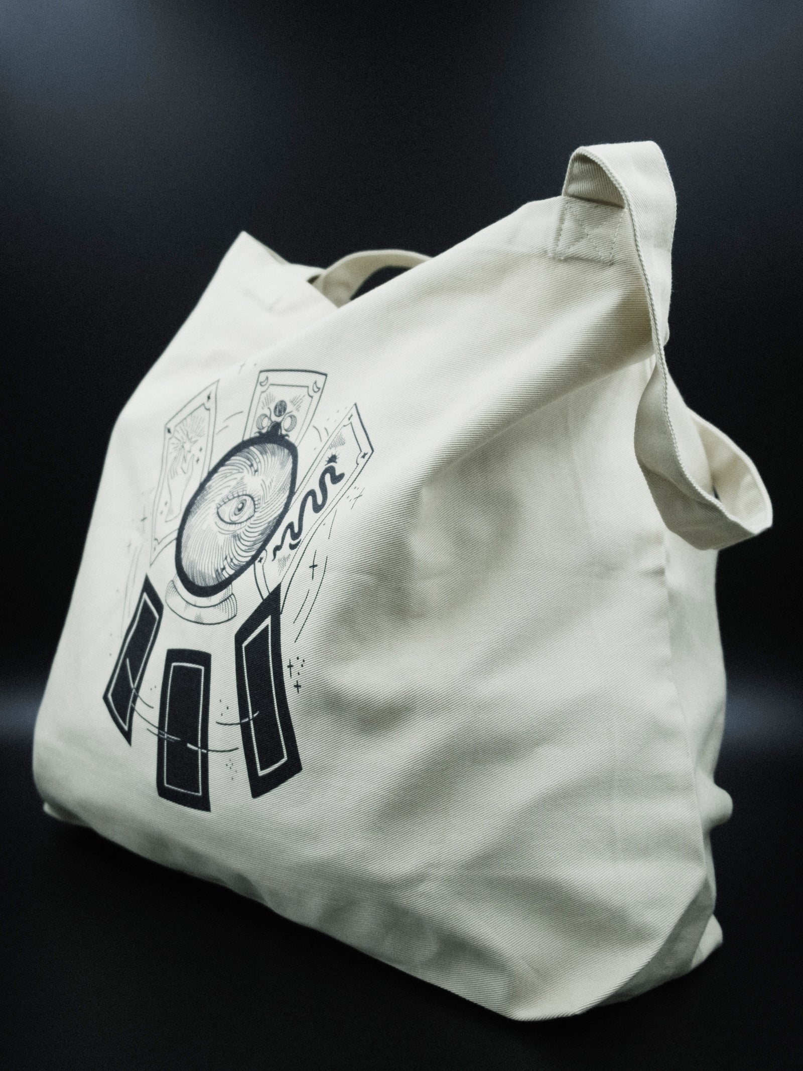 A white organic cotton tote bag with dual straps and a black graphic of a crystal ball and tarot cards on the front, placed on a dark surface.