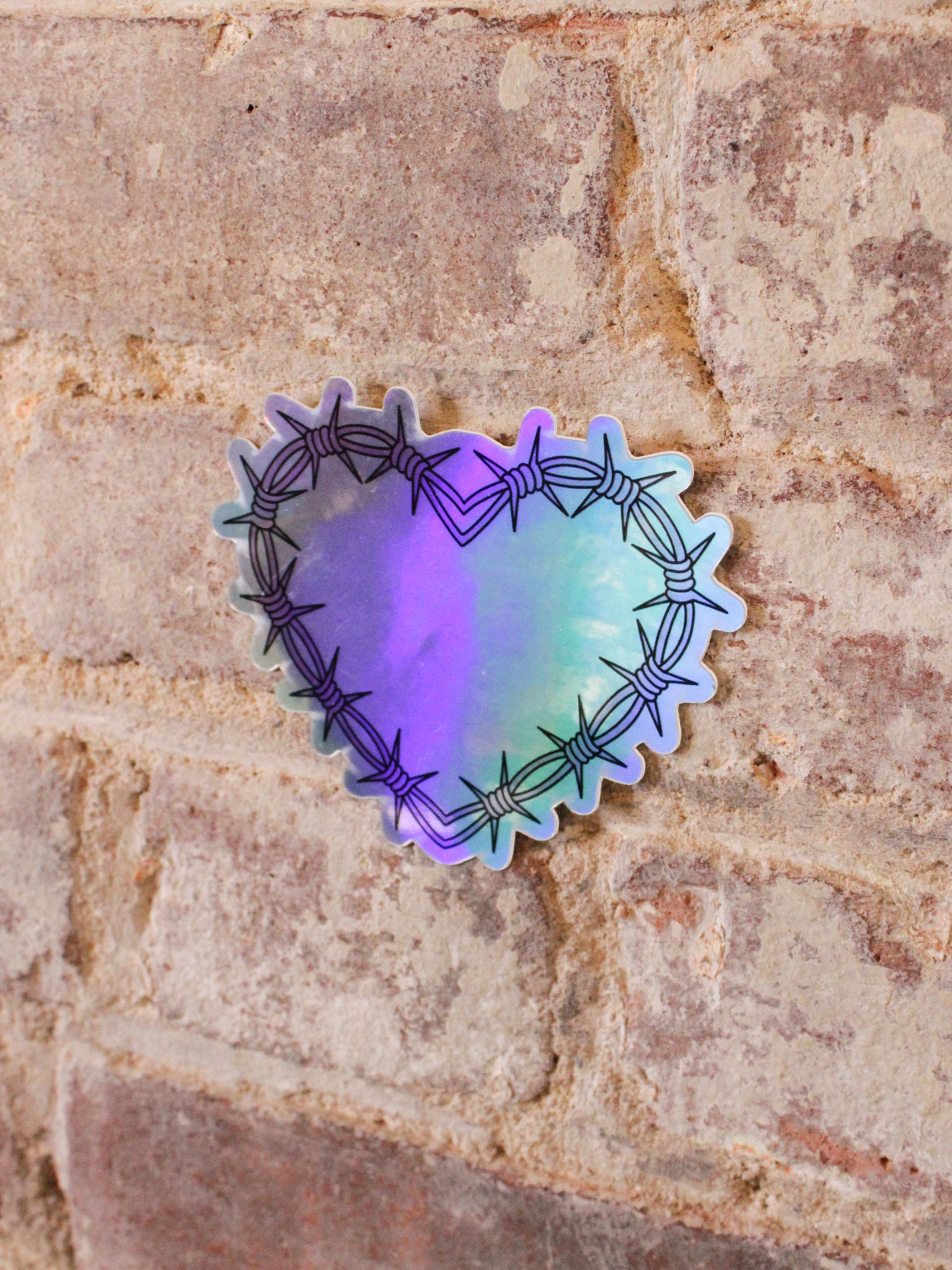 A heart-shaped holographic vinyl sticker with a pink barbed wire design in pink and black, placed on a brick surface.