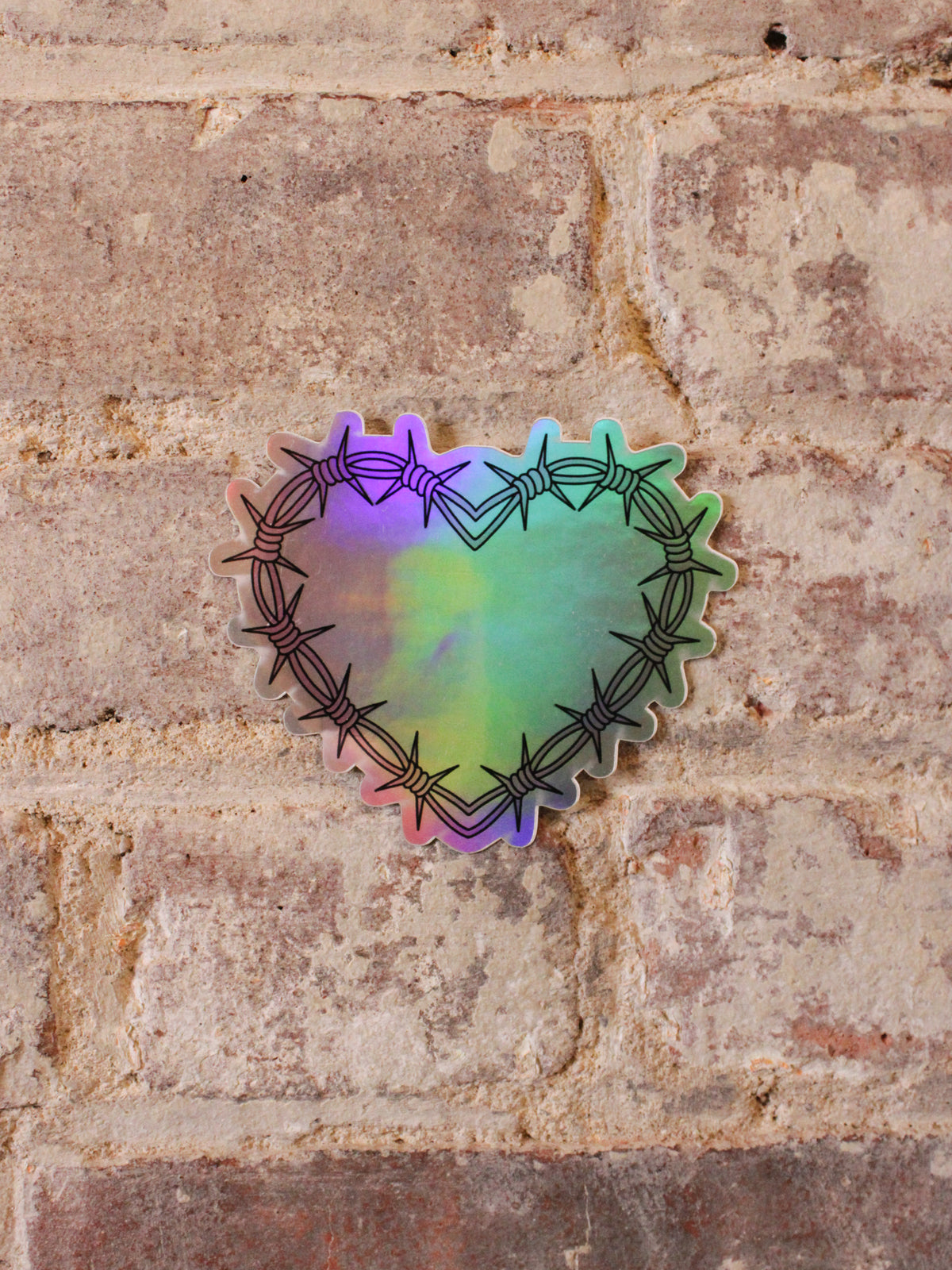 A heart-shaped holographic vinyl sticker with a pink barbed wire design in pink and black, placed on a brick surface.