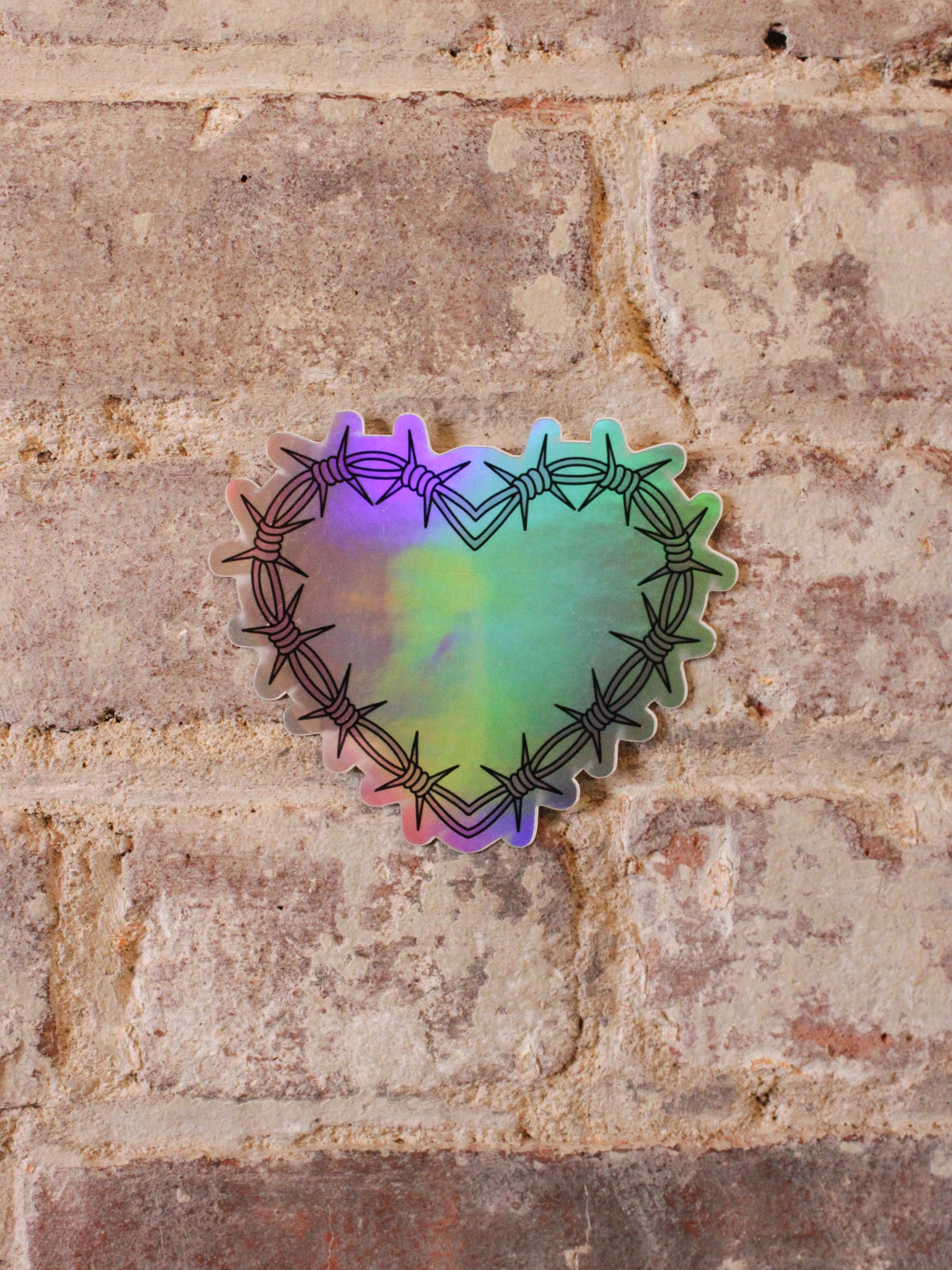 A heart-shaped holographic vinyl sticker with a pink barbed wire design in pink and black, placed on a brick surface.