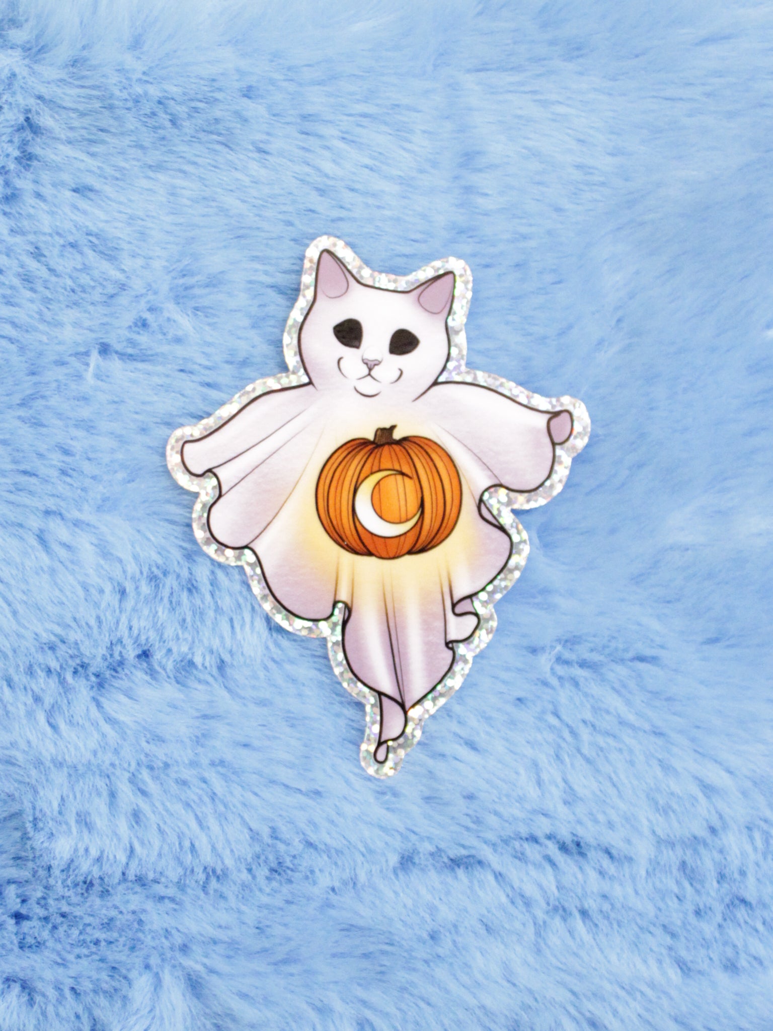 A die cut sticker of a ghost cat with a pumpkin design, with a glitter edge, placed on a blue fuzzy surface.