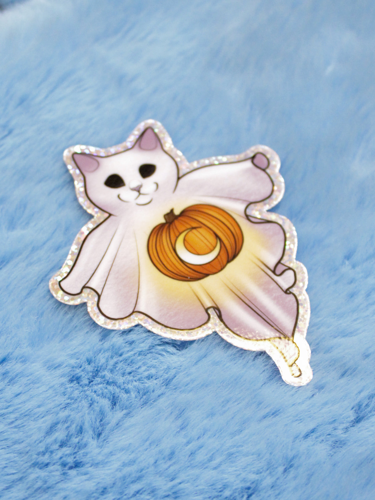 A die cut sticker of a ghost cat with a pumpkin design, with a glitter edge, placed on a blue fuzzy surface.
