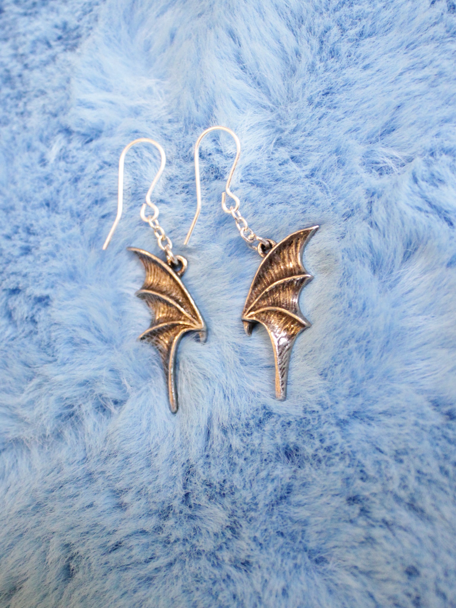 A pair of pewter and stainless steel bat-shaped dropper earrings displayed on a blue furry surface.