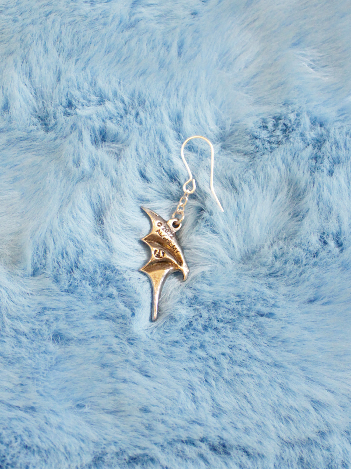 A pewter and stainless steel bat-shaped dropper earring displayed on a blue furry surface.