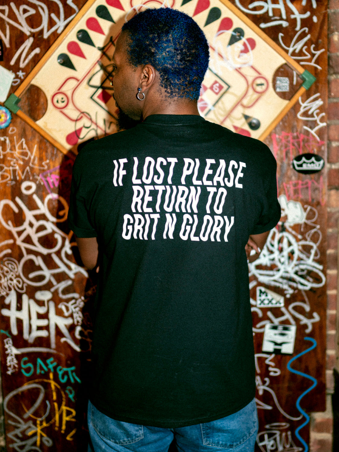 A person wearing a black graphic tee with the text 'IF LOST PLEASE RETURN TO GRIT N GLORY' on the back.