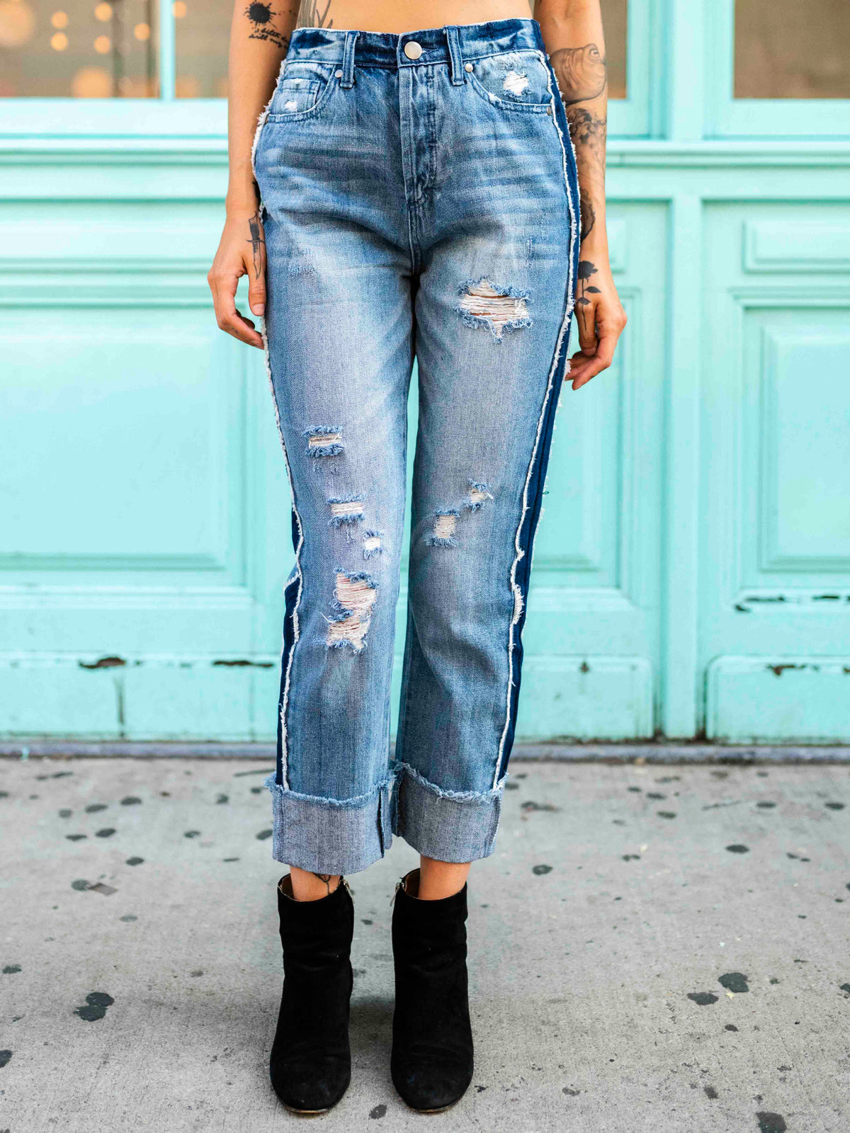 A person wearing a black crop top and blue, two-toned rolled boyfriend jeans with a frayed hem, standing in front of a teal-colored building.