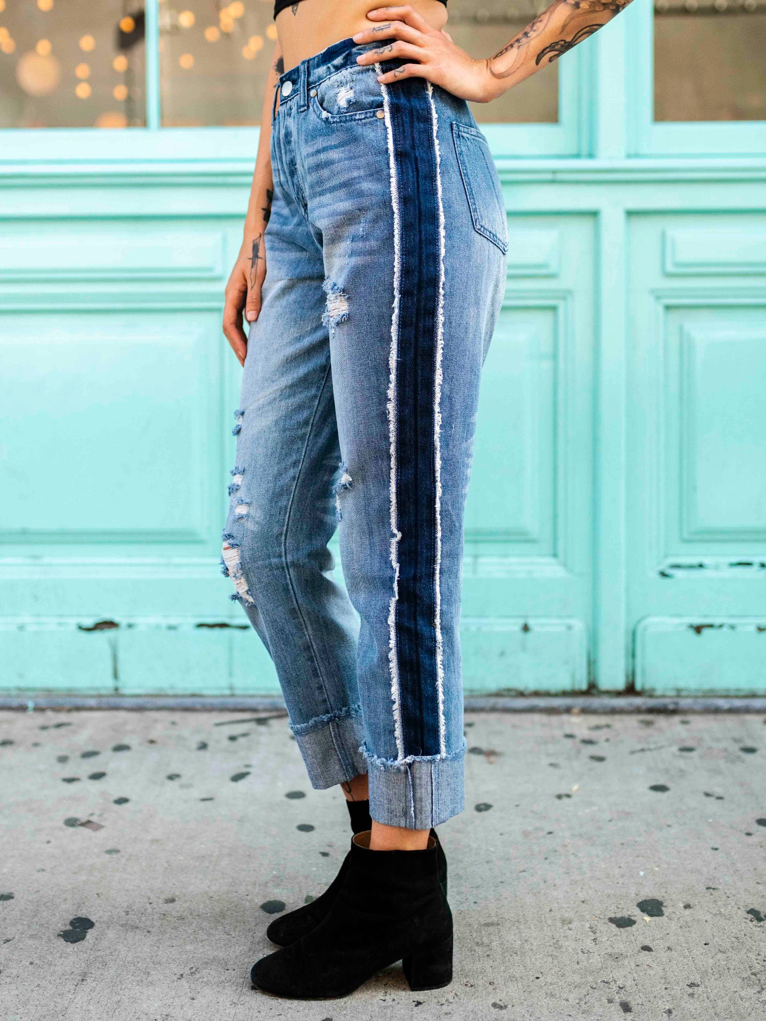 A person wearing a black crop top and blue, two-toned rolled boyfriend jeans with a frayed hem, standing in front of a teal-colored building.