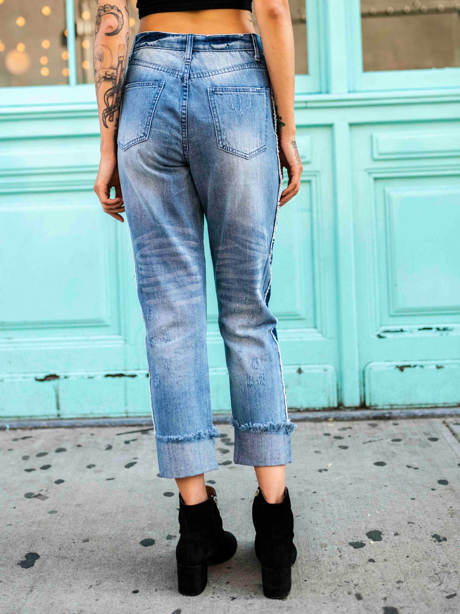 A person wearing a black crop top and blue, two-toned rolled boyfriend jeans with a frayed hem, standing in front of a teal-colored building.