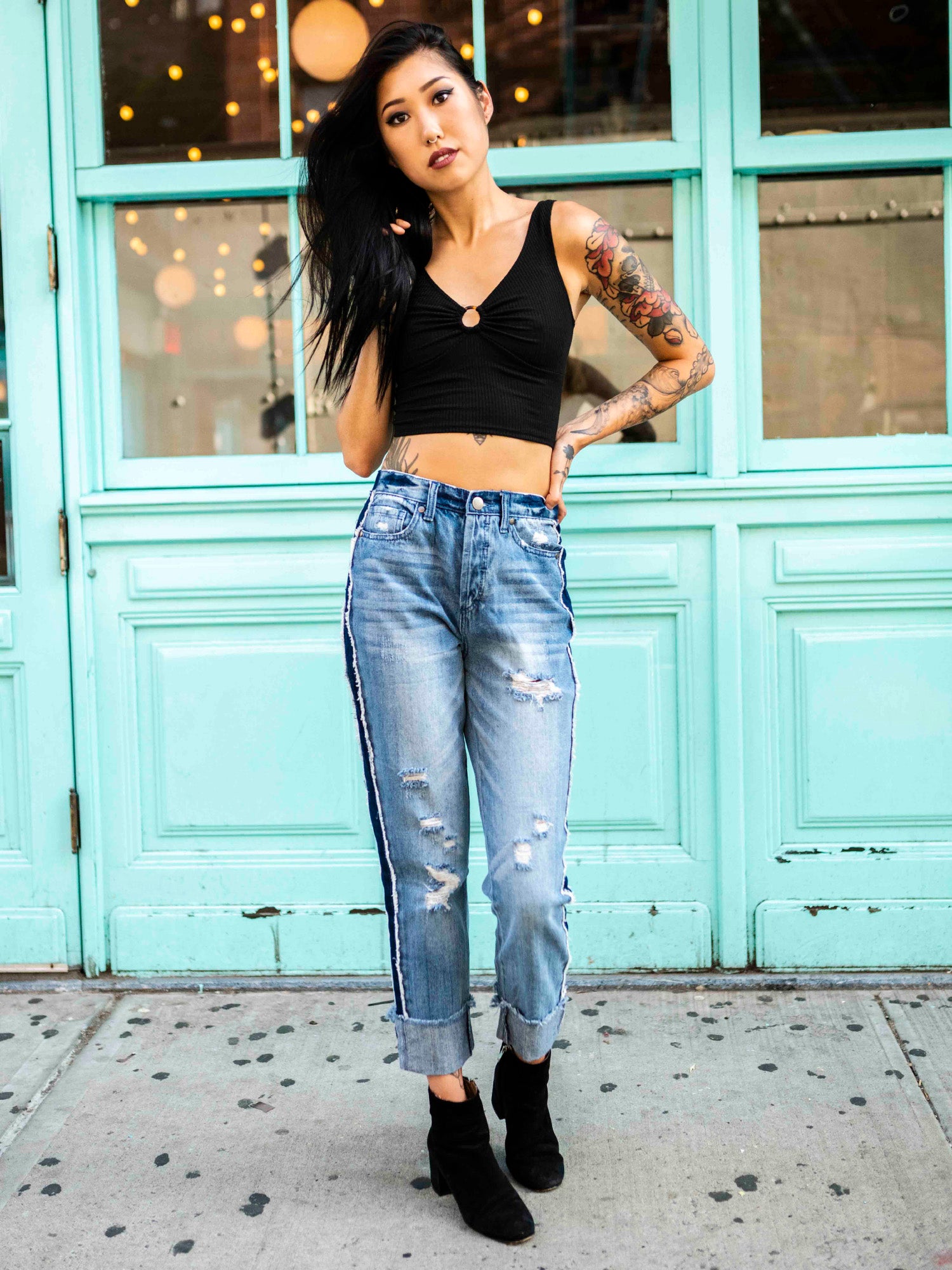 A person wearing a black crop top and blue, two-toned rolled boyfriend jeans with a frayed hem, standing in front of a teal-colored building.