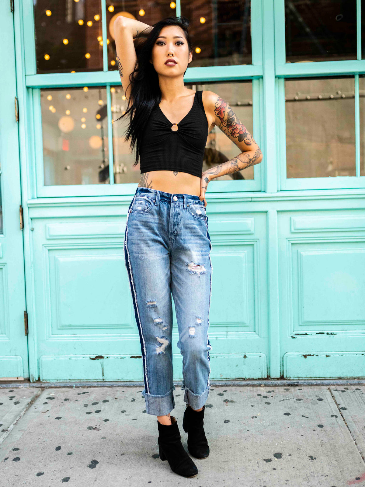 A tattooed person wearing a black crop top and blue, two-toned rolled boyfriend jeans with a frayed hem, standing in front of a teal-colored building.