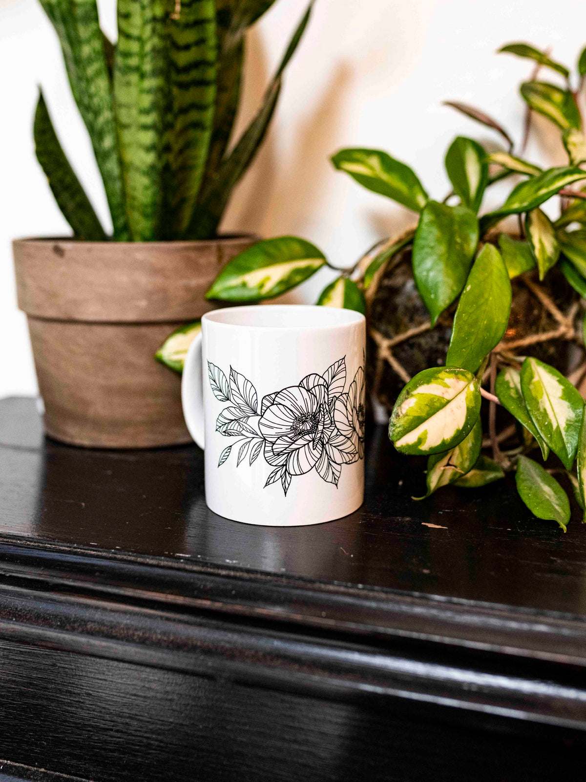 A white ceramic mug with a black floral pattern designed by tattoo artist Janice Danger, placed on a dark surface with green plants in the background.