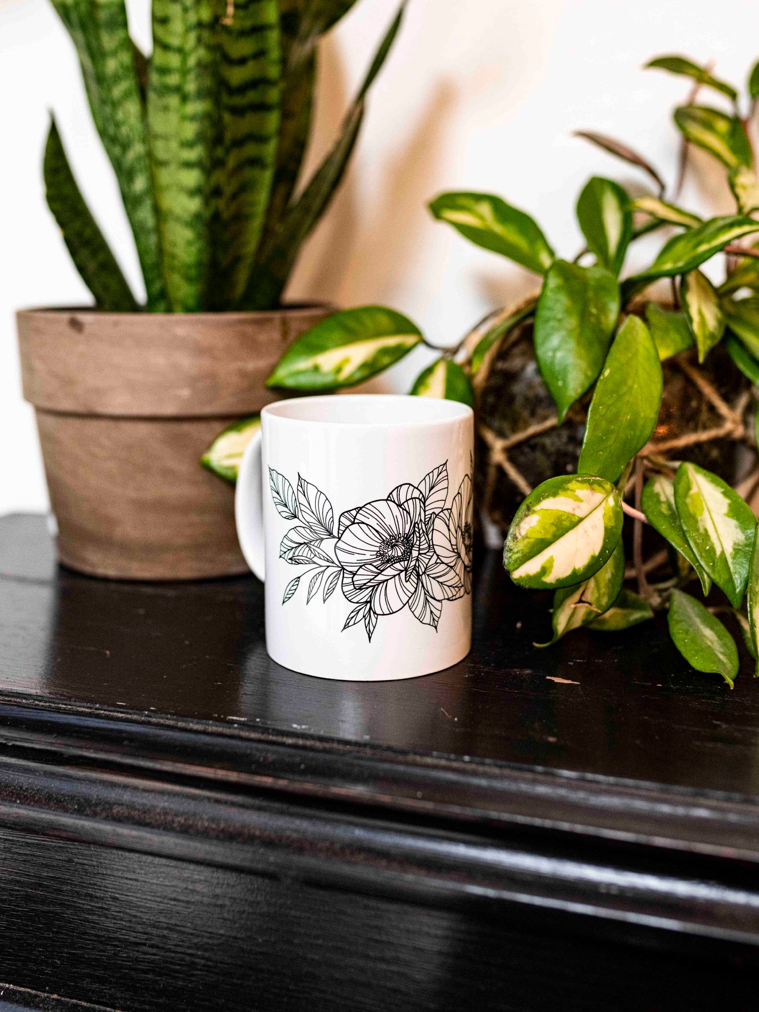 A white ceramic mug with a black floral pattern designed by tattoo artist Janice Danger, placed on a dark surface with green plants in the background.