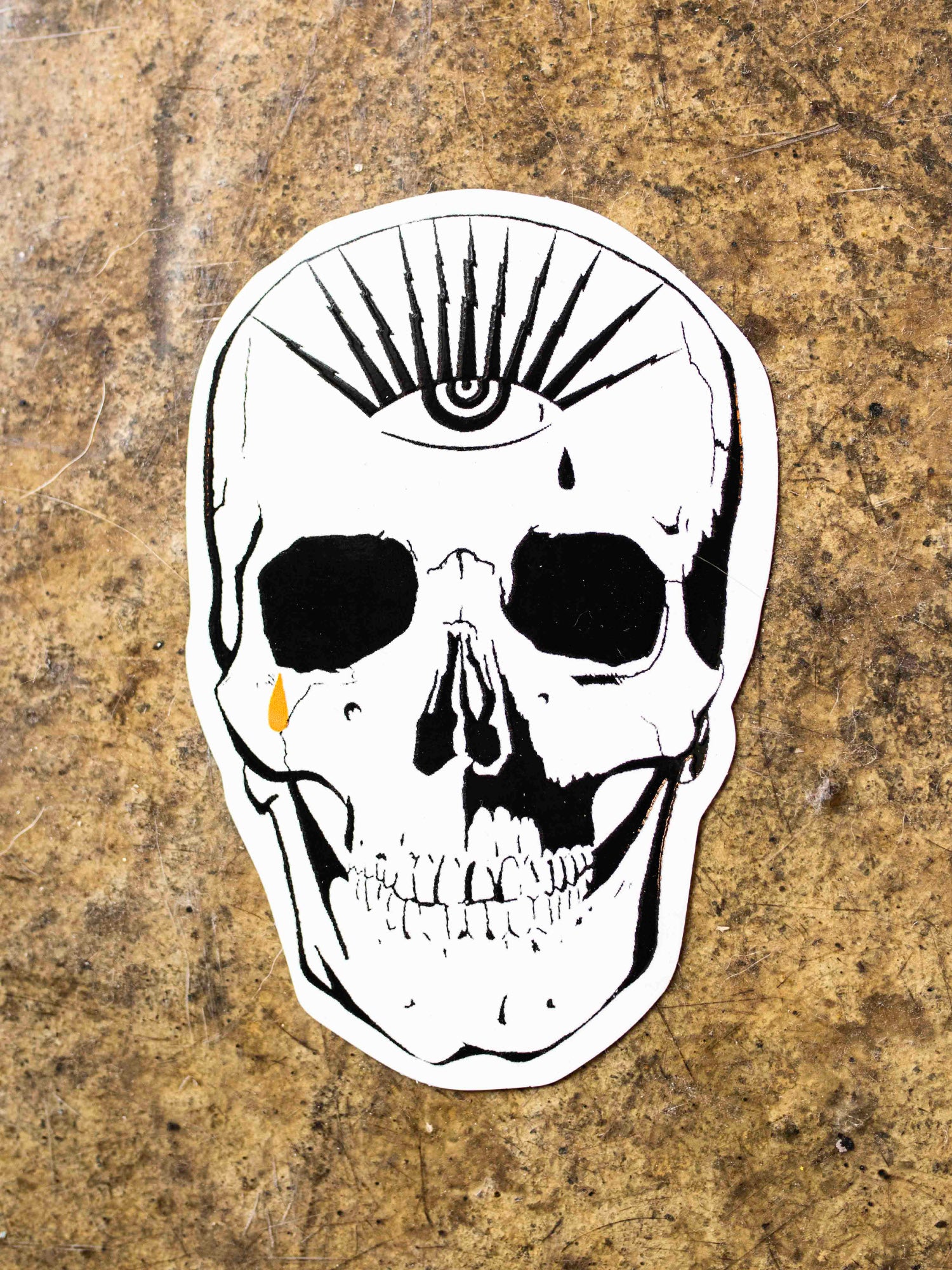 A black, white, and gold die-cut sticker of a skull with an eye the top forehead area.