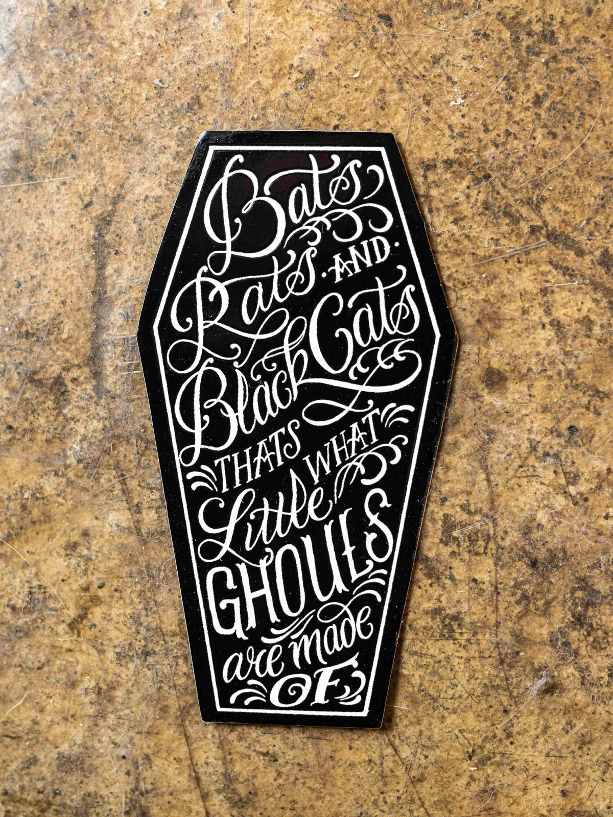 A sticker with text designed by Megan Massacre placed on a concrete background. Sticker is in a coffin shape and reads "Bats, rats and black cats that's what little ghouls are made of"
