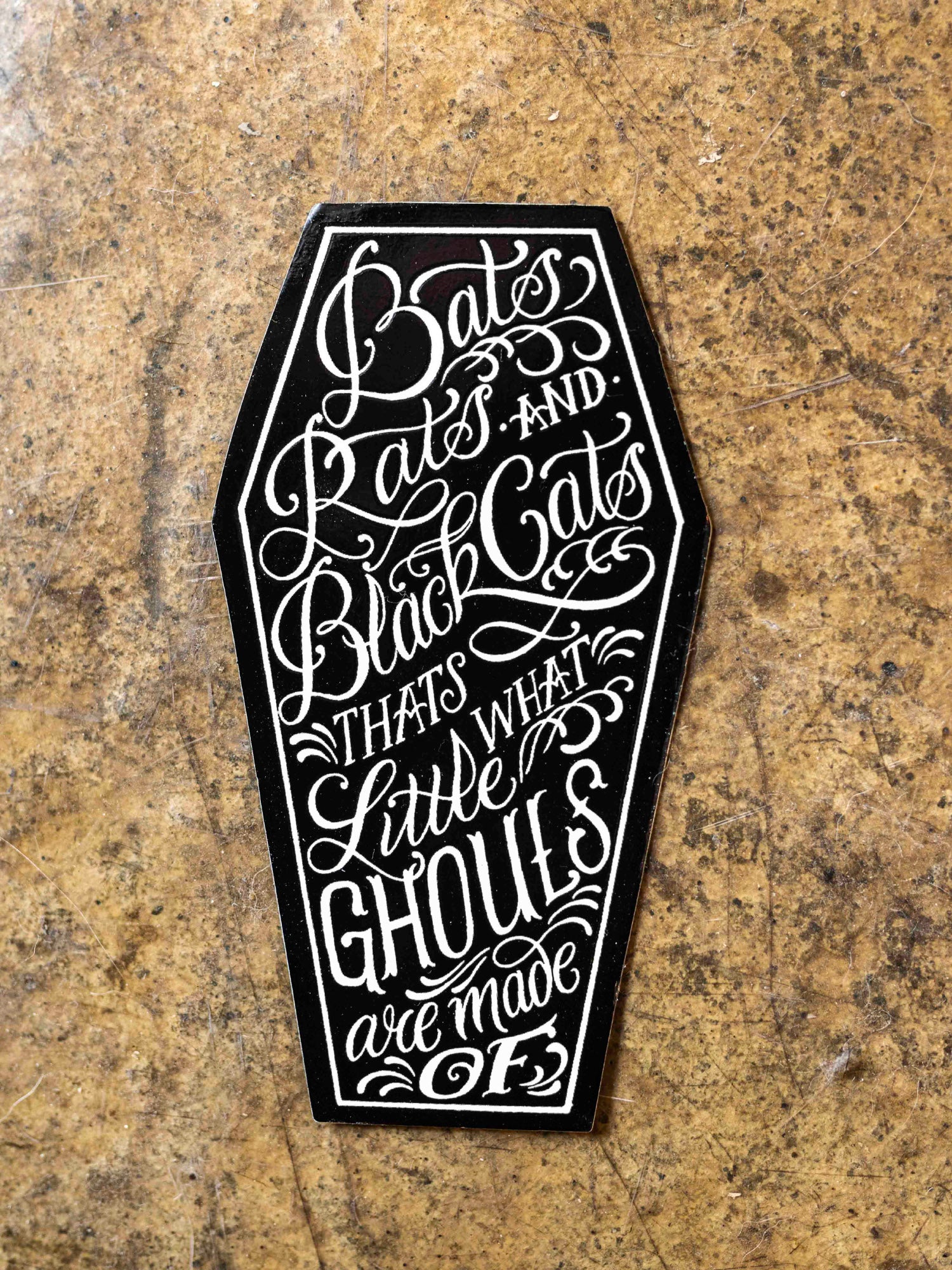 A sticker with text designed by Megan Massacre placed on a concrete background. Sticker is in a coffin shape and reads "Bats, rats and black cats that's what little ghouls are made of"