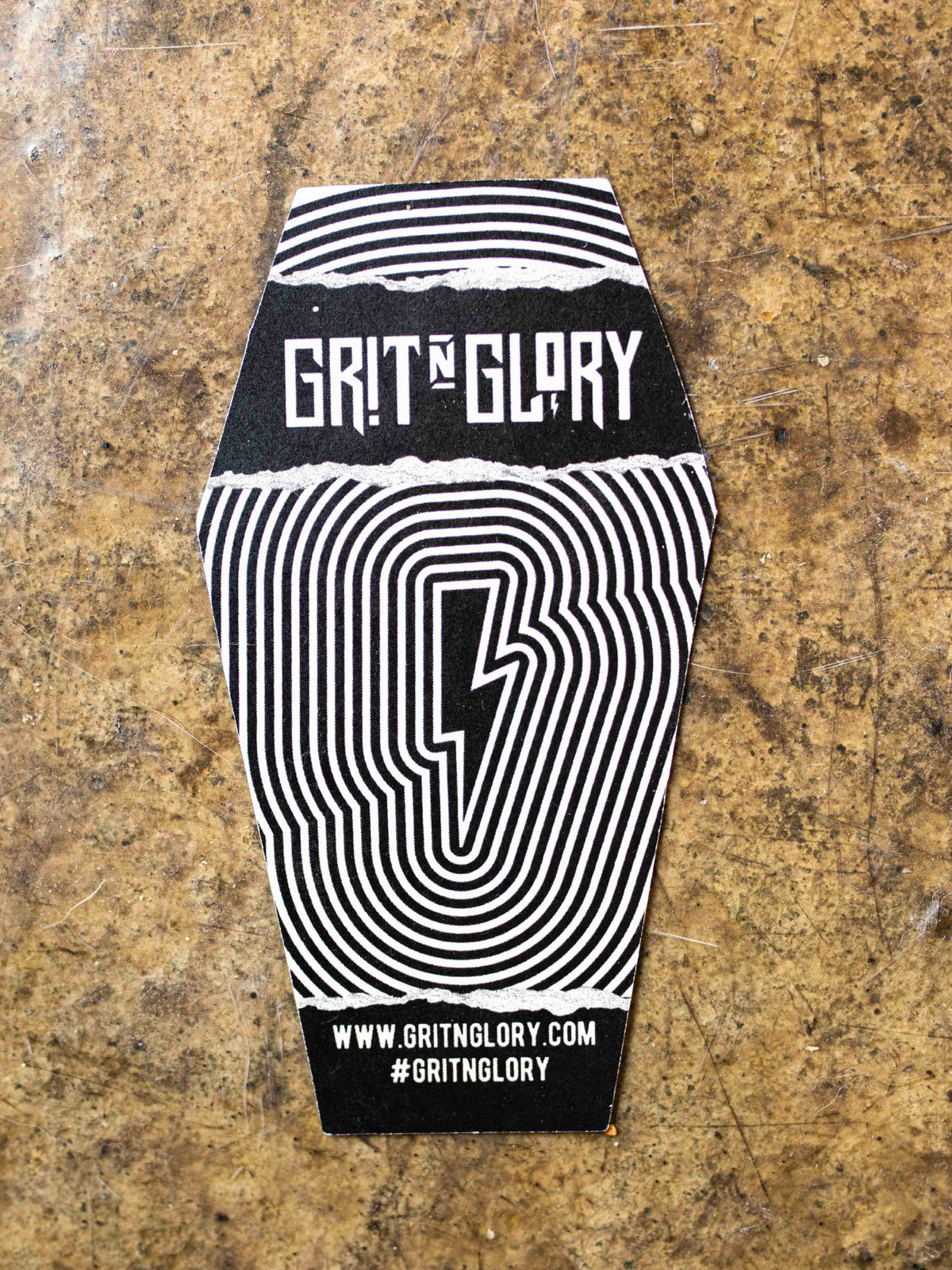 The back of a sticker with text designed by Megan Massacre placed on a concrete background. Sticker is in a coffin shape and reads "Bats, rats and black cats that's what little ghouls are made of"