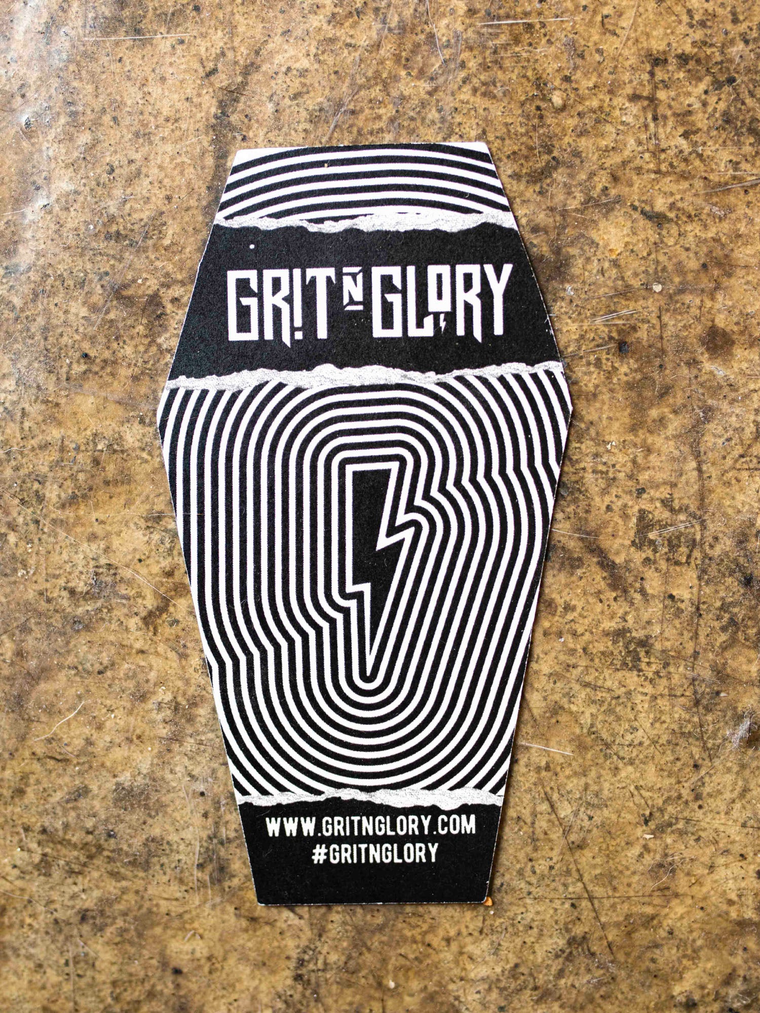 The back of a sticker with text designed by Megan Massacre placed on a concrete background. Sticker is in a coffin shape and reads "Bats, rats and black cats that's what little ghouls are made of"