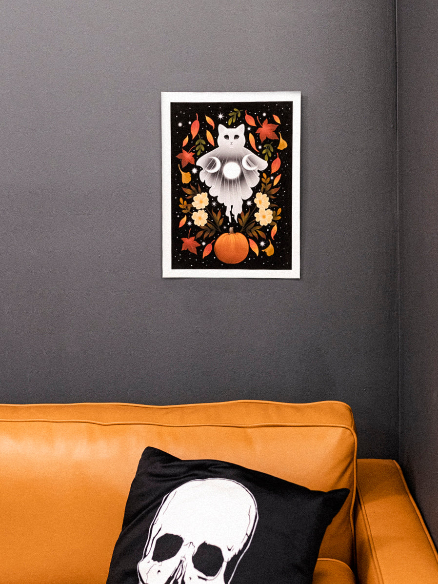 Autumn Cat Ghost art print hanging on a dark wall, displaying a white cat ghost with moons, autumn leaves, a pumpkin and florals on a black starry background. 