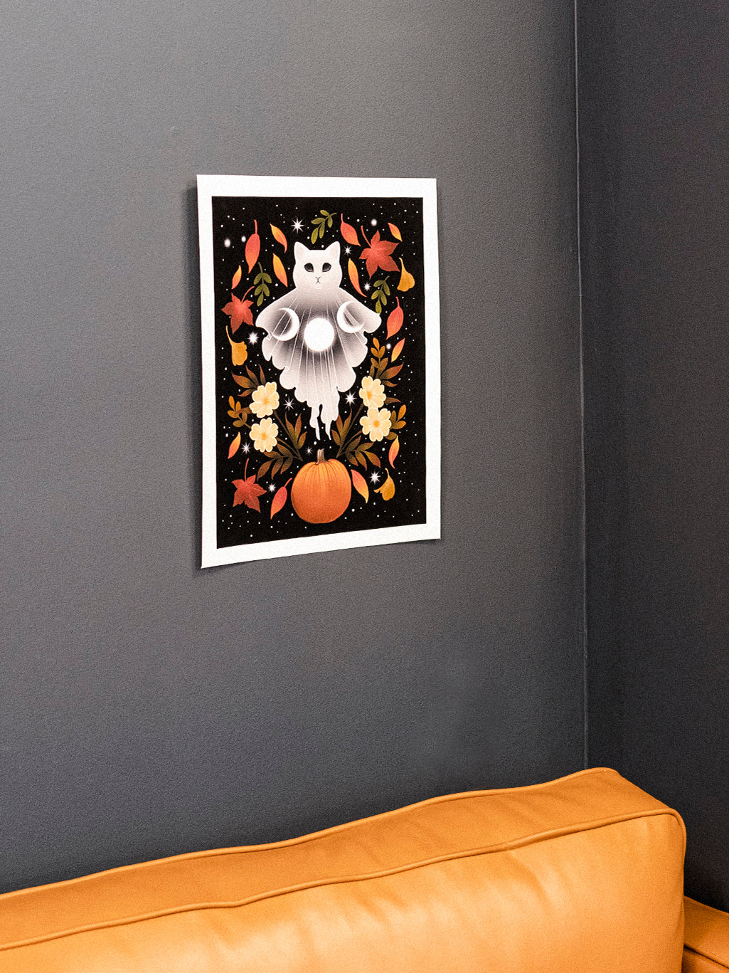 Autumn Cat Ghost art print hanging on a dark wall above an orange sofa, displaying a white cat surrounded by fall-themed decorations.