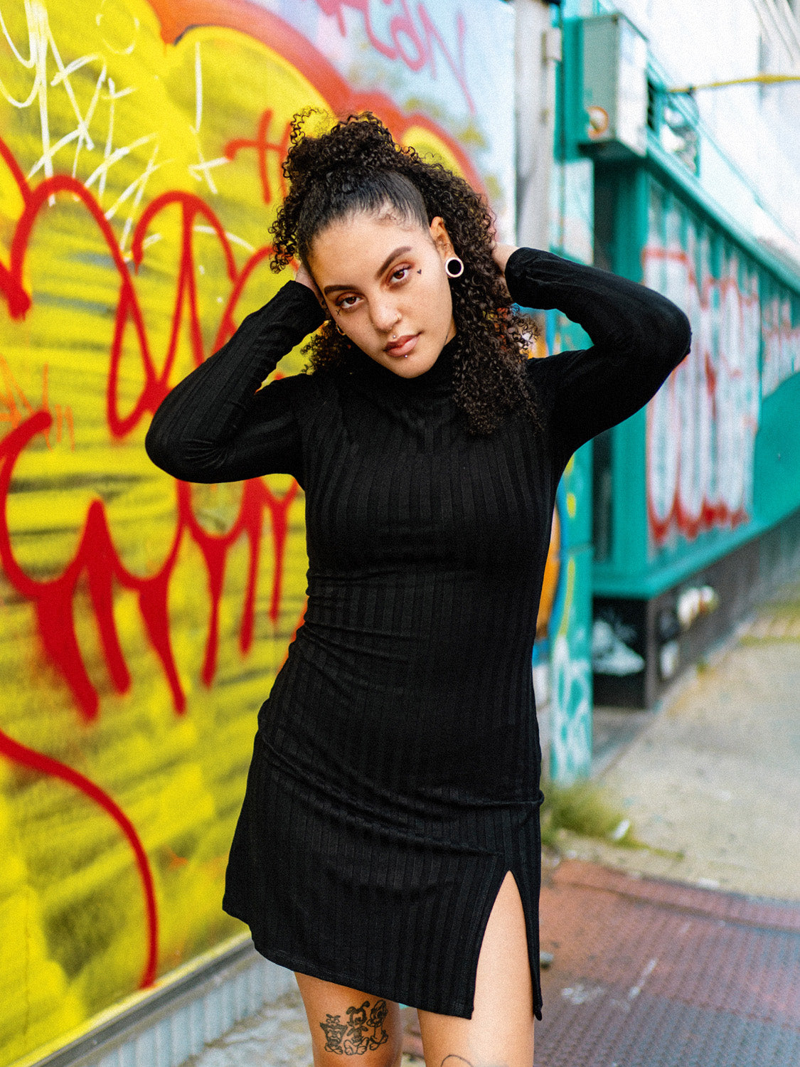 A person wearing a black mini dress with a mock neck, long sleeves, and a side slit. The dress has a bodycon fit and the person is paired with black boots, standing in front of a colorful graffiti wall.