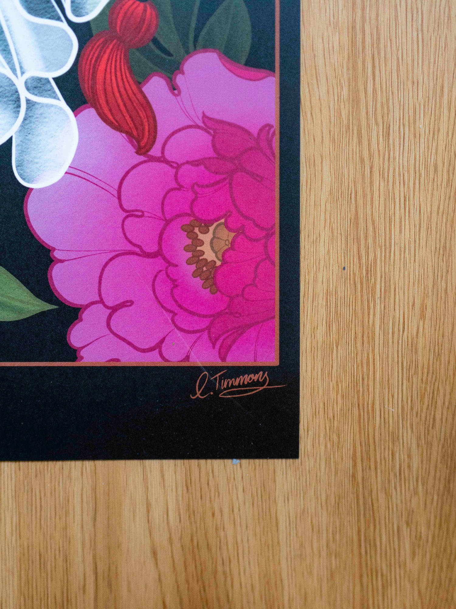 Signature detail of an art print featuring a stylized white cat figure with floral elements, displayed on a wooden table.