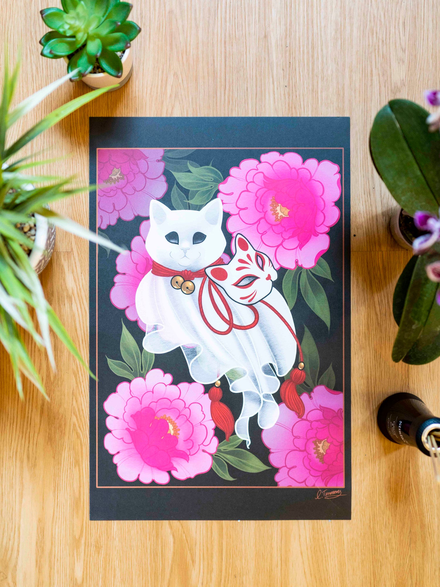 An art print featuring a stylized white cat figure with floral elements, displayed on a wooden table with potted plants.