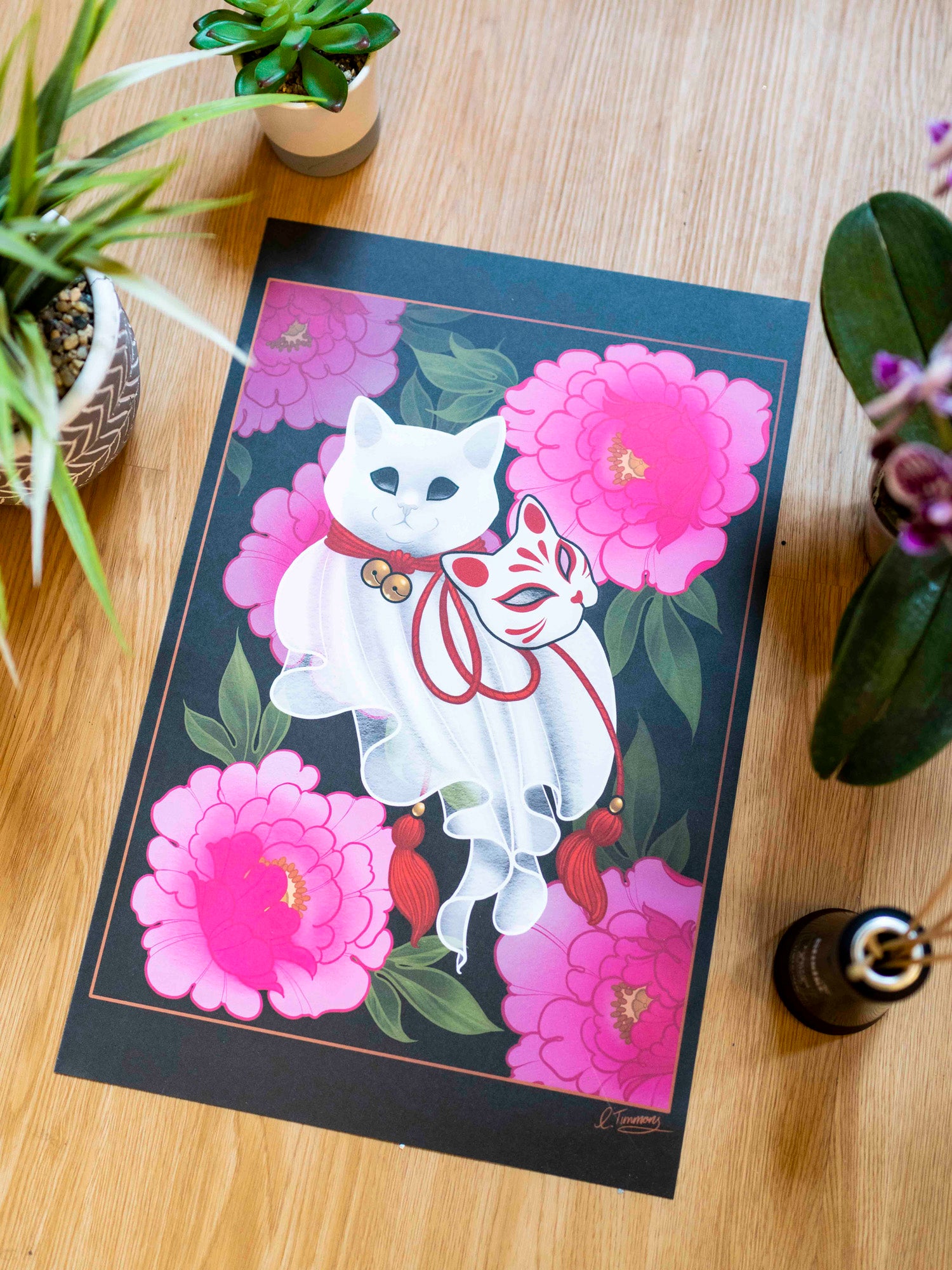 An art print featuring a stylized white cat figure with floral elements, displayed on a wooden table with potted plants.