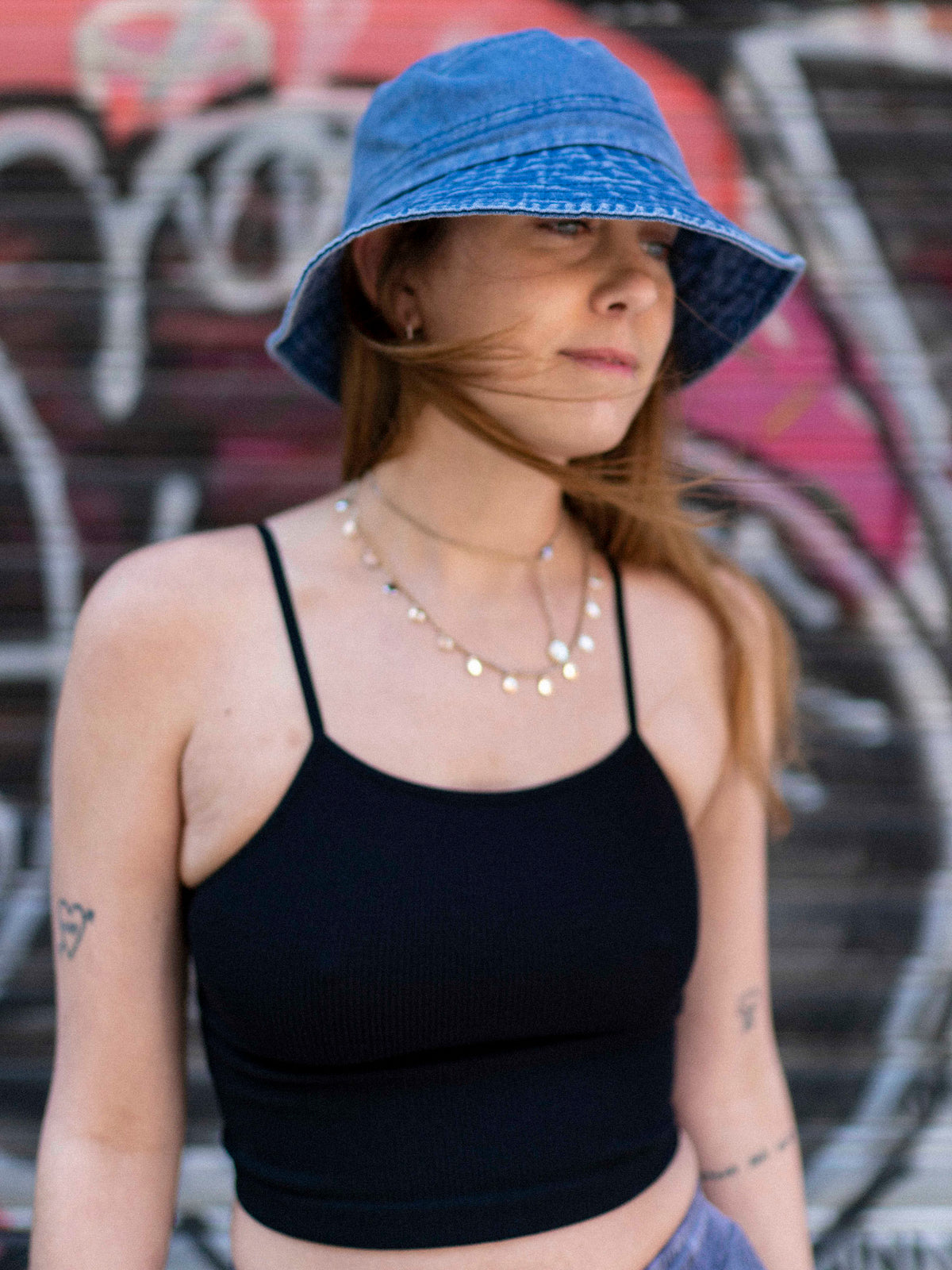 A person wearing a blue denim bucket hat and a black sleeveless top, standing in front of a graffiti-covered wall.