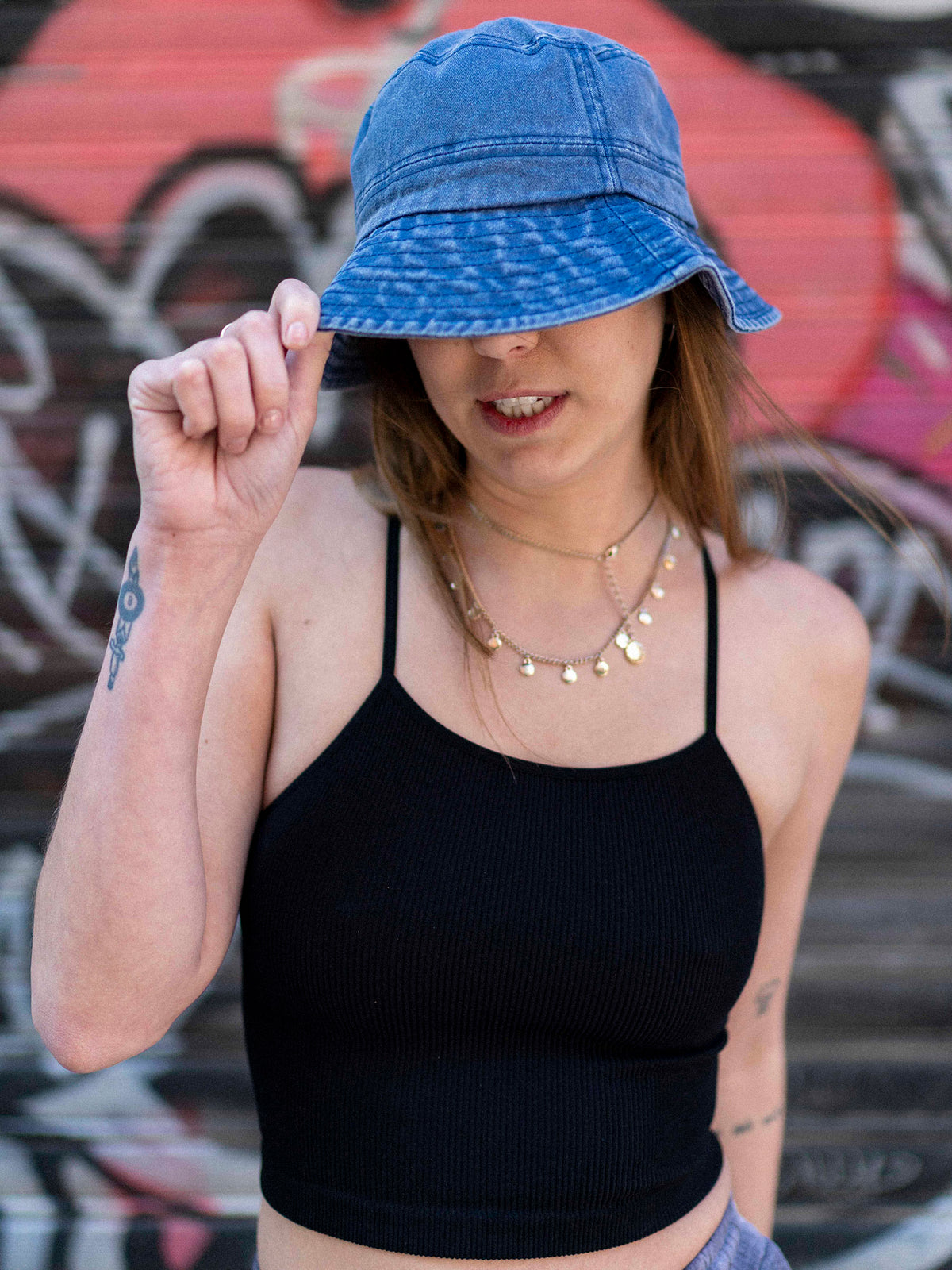 A person wearing a blue denim bucket hat and a black sleeveless top, standing in front of a graffiti-covered wall.