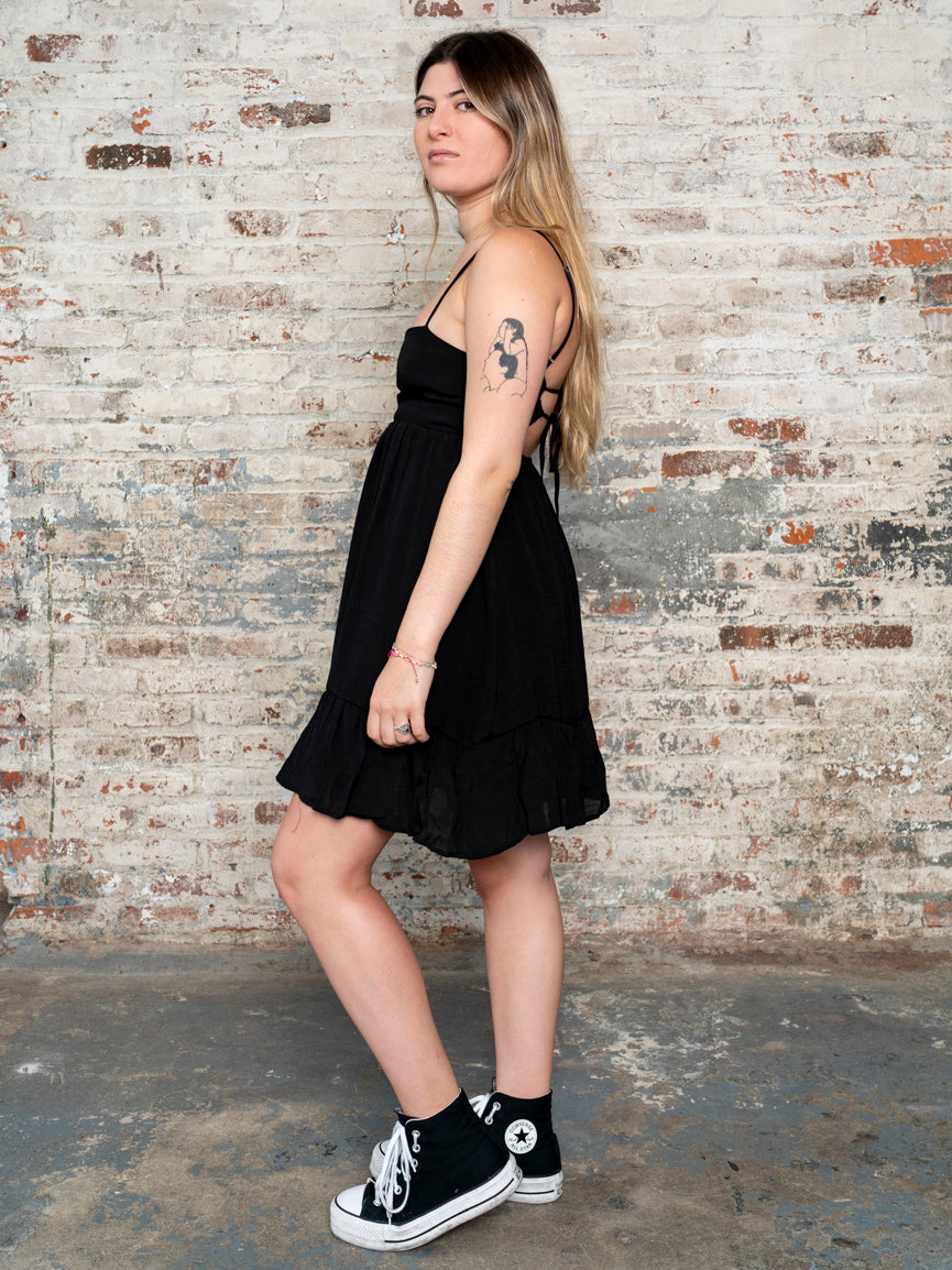 model wearing a black cami strapped dress with classic converse sneakers against a rustic brick wall. 