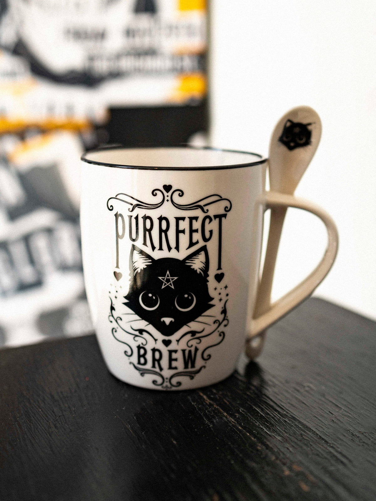 A white ceramic mug with a black cat design and the text 'Purrfect Brew' on it, accompanied by a matching white spoon with a black handle design.