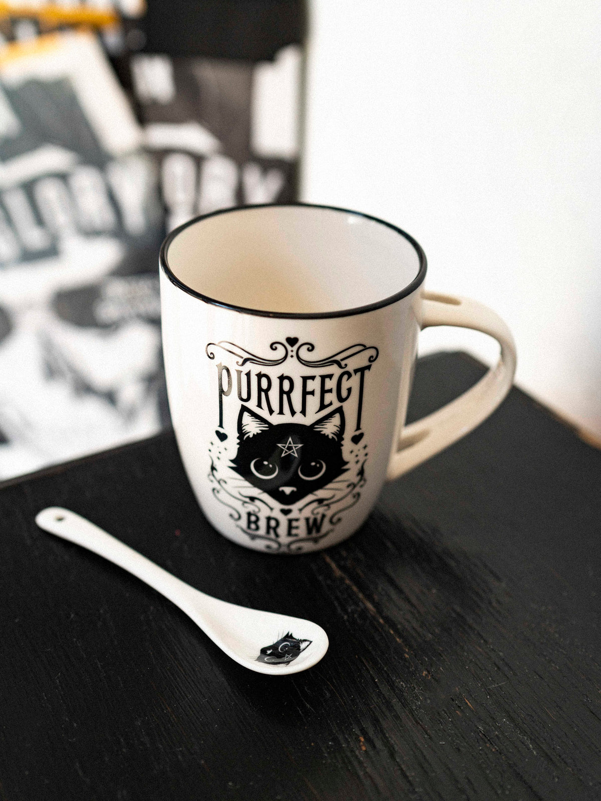 A white ceramic mug with a black cat design and the text &#39;Purrfect Brew&#39; on it, accompanied by a matching white spoon with a black handle design.
