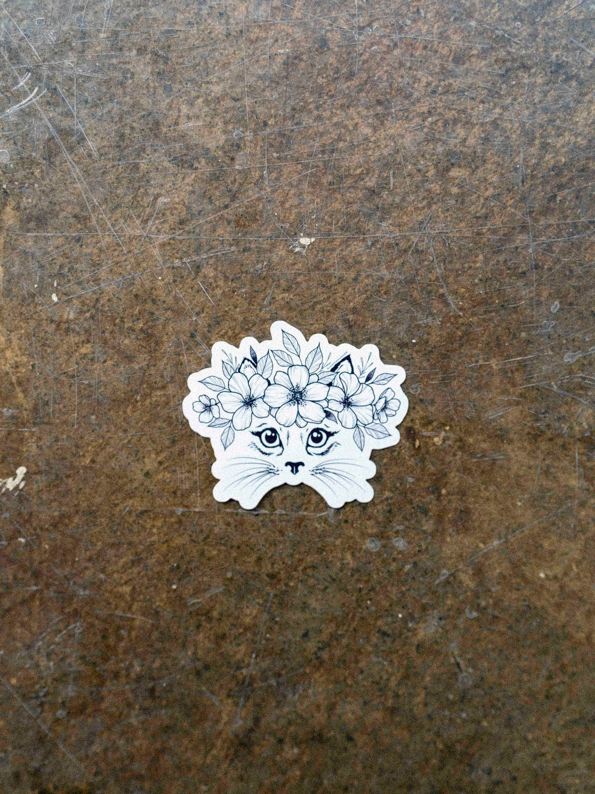 A black and white vinyl sticker of a cat with a floral crown illustration by Janice Danger on a concrete background.