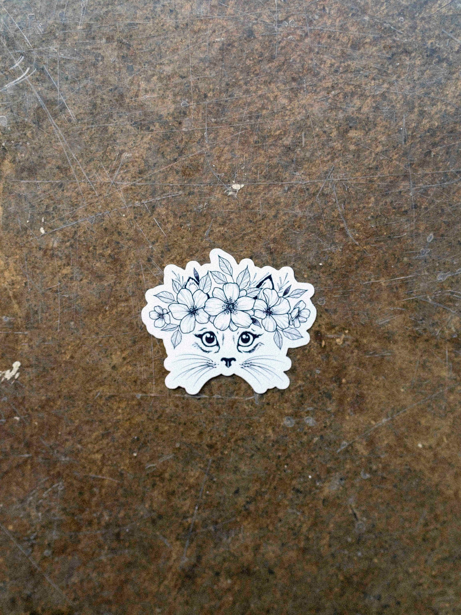 A black and white vinyl sticker of a cat with a floral crown illustration by Janice Danger on a concrete background.