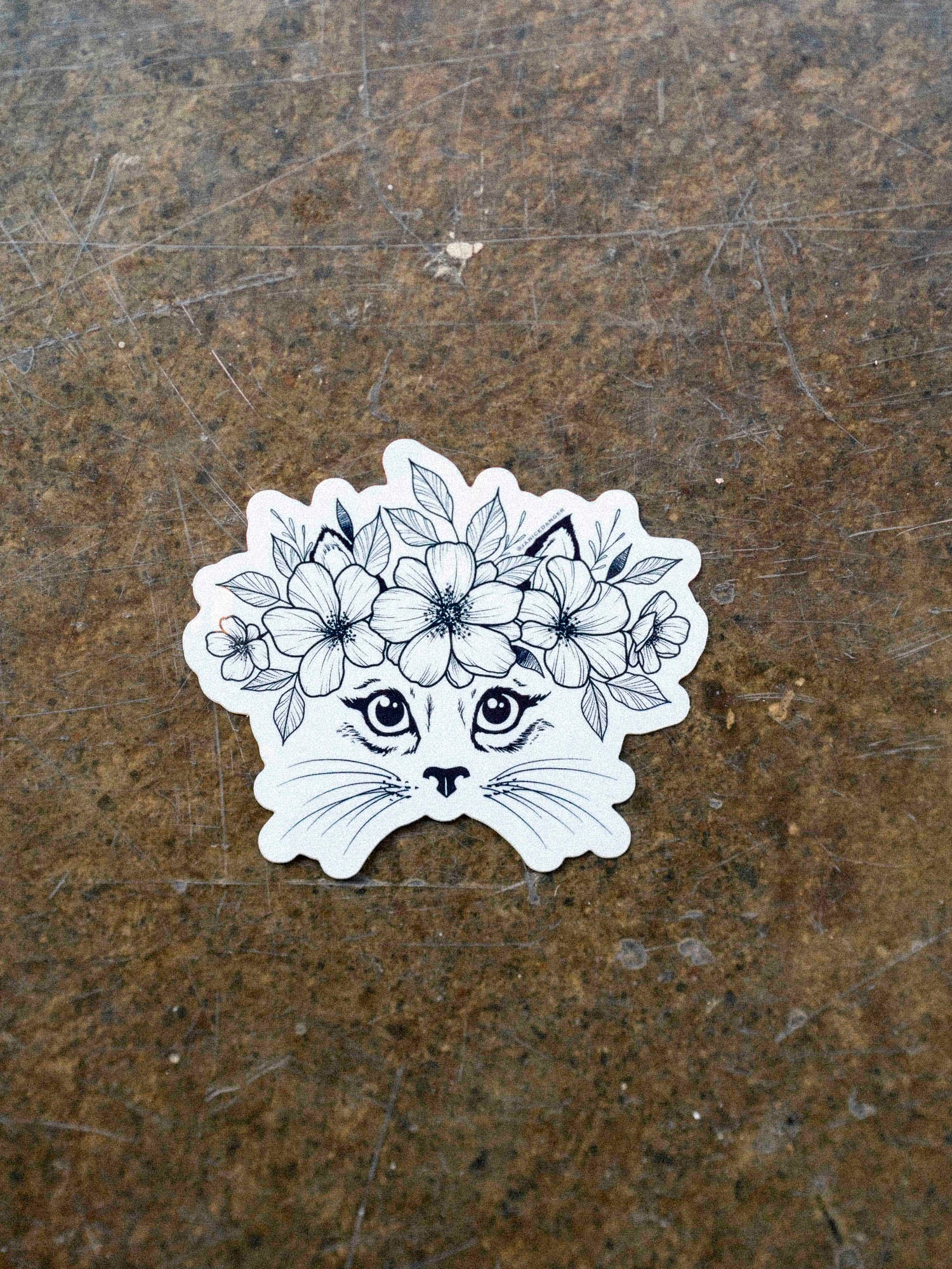A black and white vinyl sticker of a cat with a floral crown illustration by Janice Danger on a concrete background.